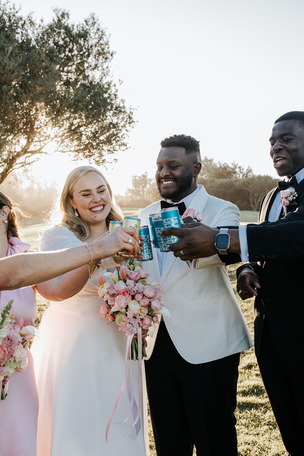 Bride and groom toasting with canned drinks alongside wedding guests outdoors in golden hour sunlight.