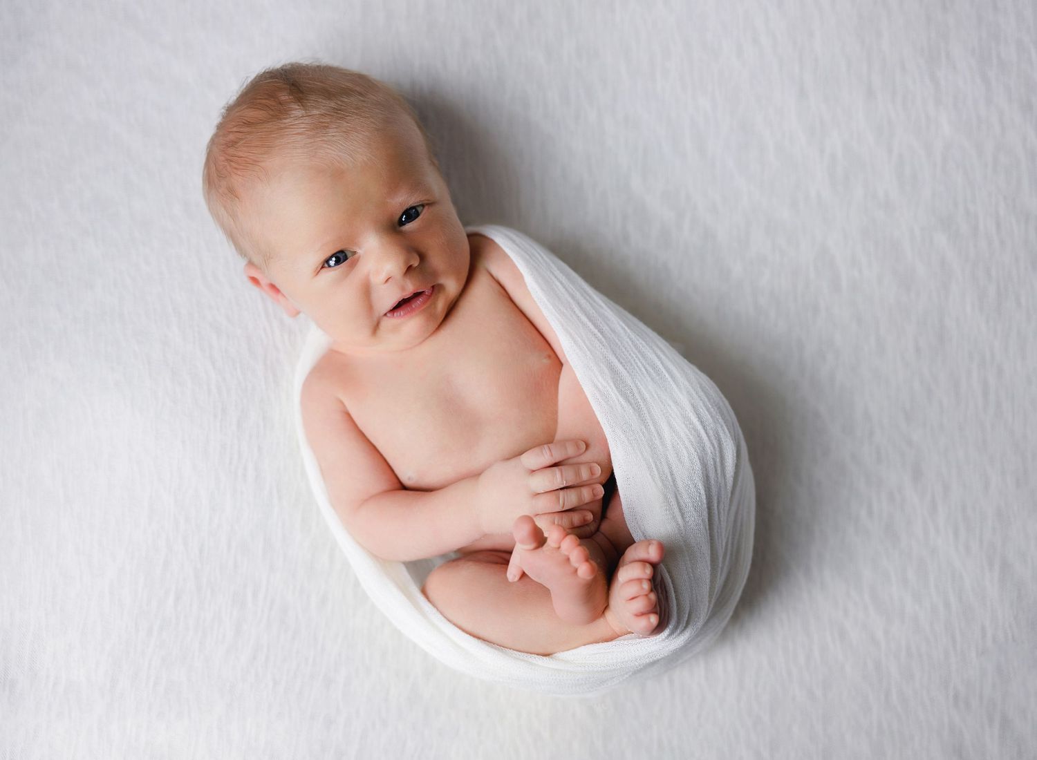 A tiny baby wrapped in soft white fabric lies on a white backdrop while smiling sweetly.