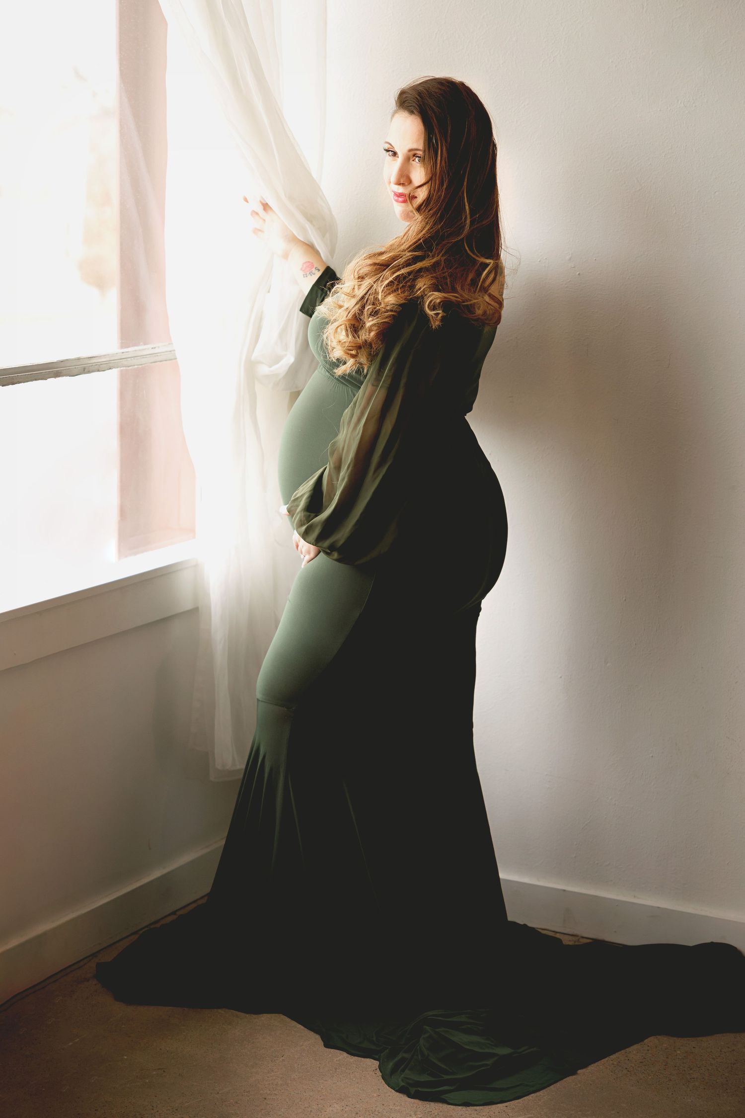 Elegant maternity silhouette in green dress stands by window with flowing curtains in natural light.