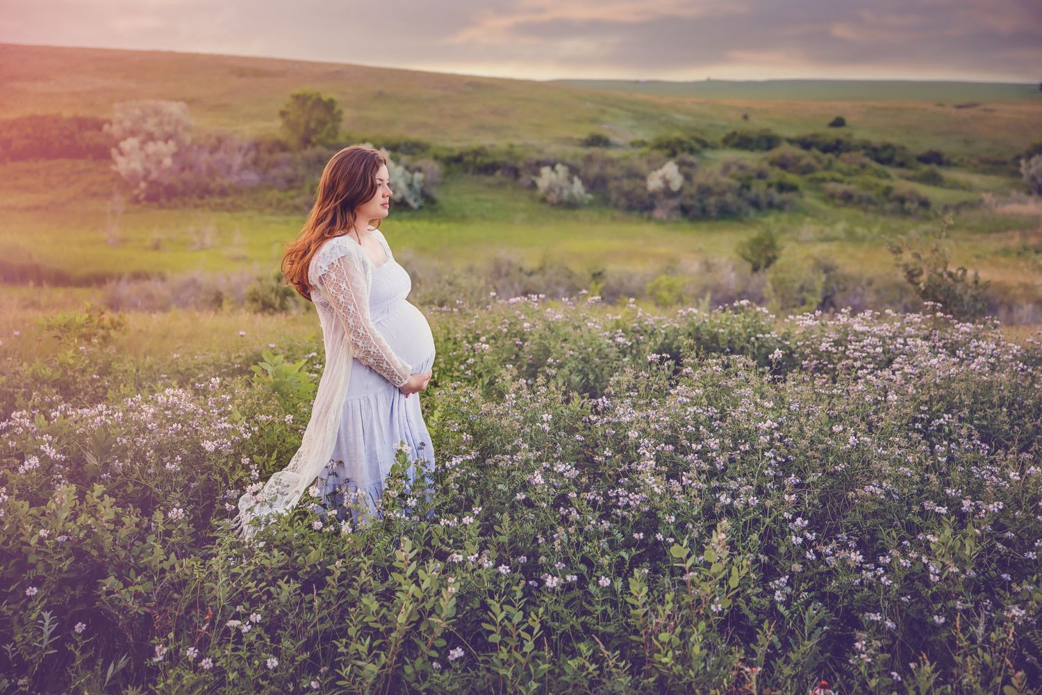 Maternity Woman in flowing white dress stands among wildflowers in sunset meadow.