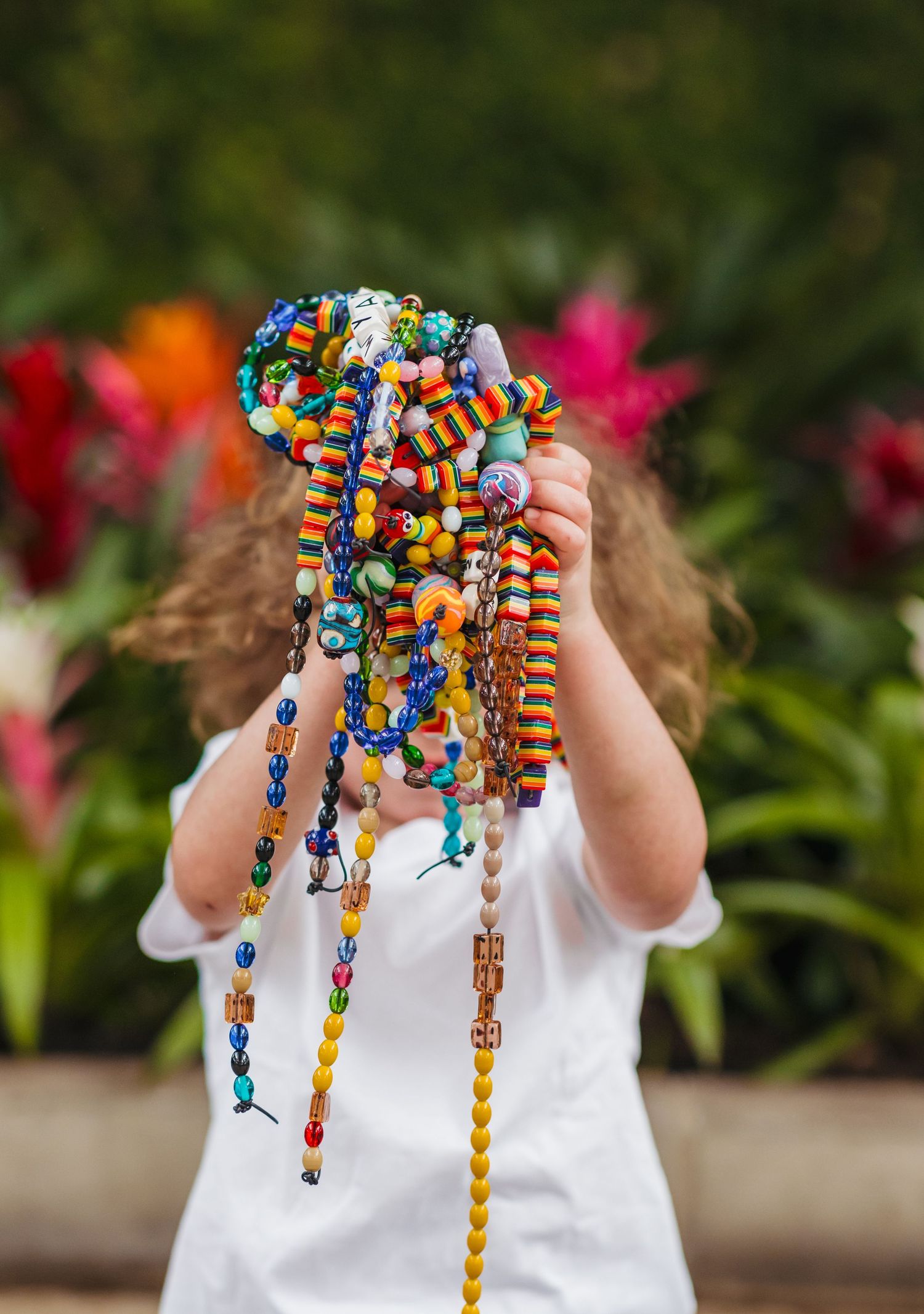 Child holding colorful beaded necklaces and bracelets in front of face with vibrant flowers in background.