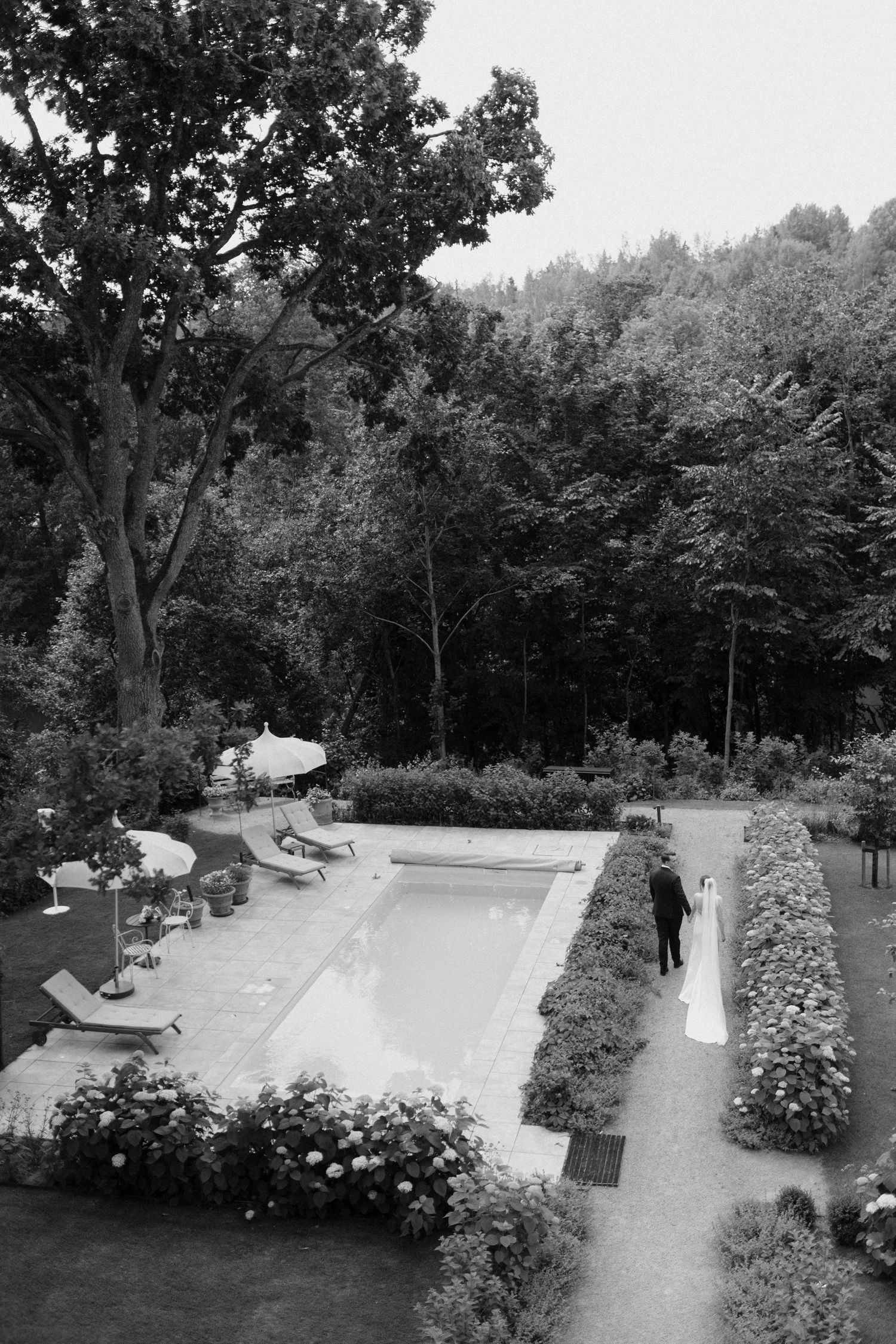 An editorial wedding portrait of a bride and groom walking hand in hand by the pool in a garden in Billnäs Gård, Finland.