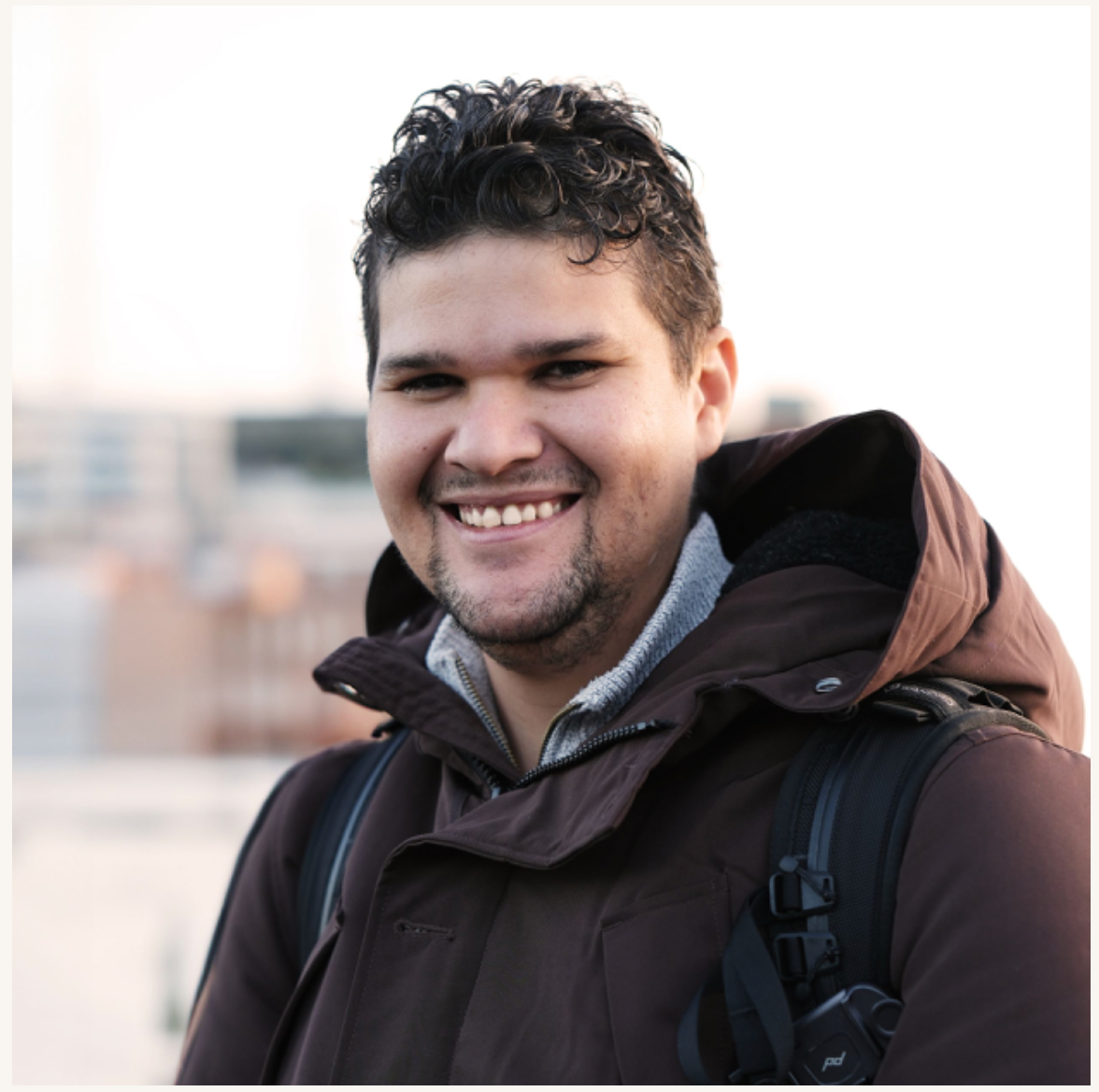 Person in brown winter coat smiling warmly at camera while wearing backpack outdoors.