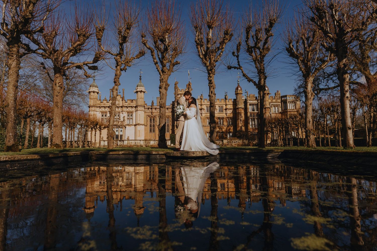 A couple in wedding attire stands reflected in a pond with bare winter trees and a grand mansion in the background.