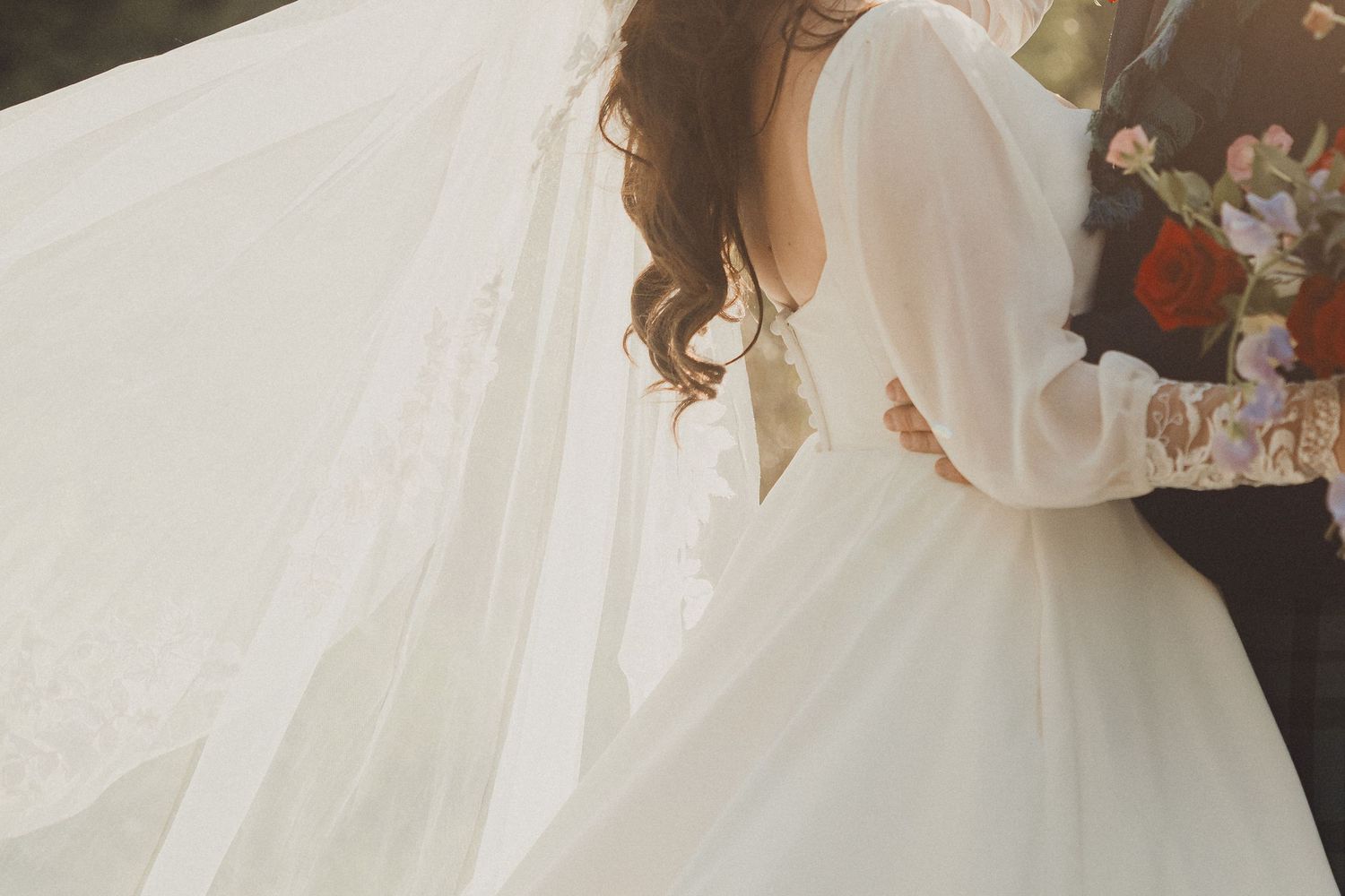 Bride in elegant white wedding gown with flowing veil embracing groom outdoors in soft natural light.