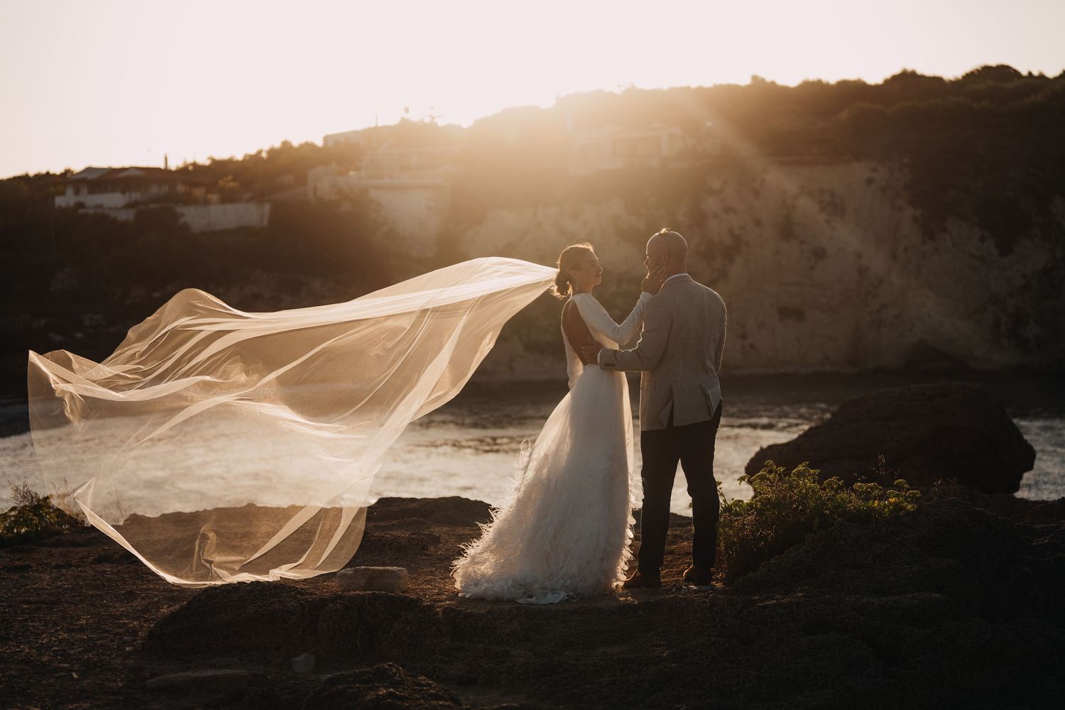 A couple embraces at sunset while a long wedding veil flows dramatically in the wind on a hilltop.