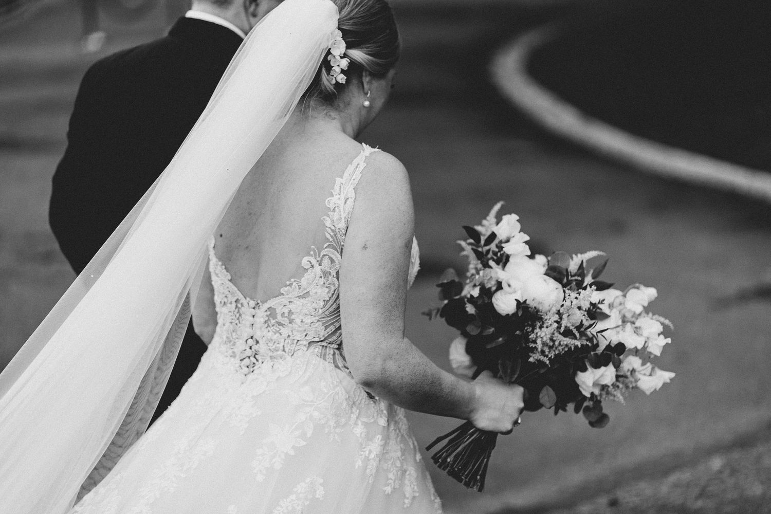 Black and white photo of bride in lace dress holding bouquet with flowing veil.