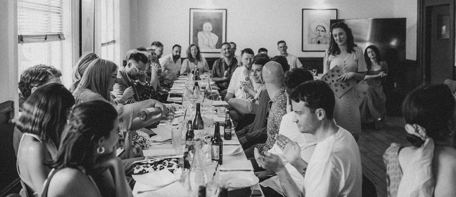 Black and white photo of a casual wedding dinner party at the Camberwell Arms in London, guests sit at either side of a long table chatting