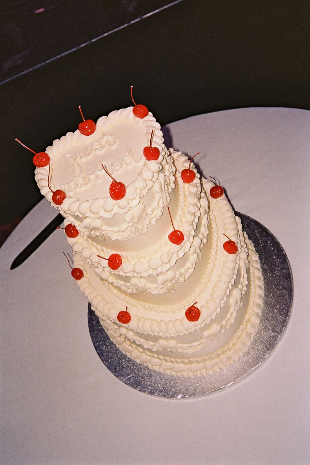 Three-tier retro-style cream wedding cake decorated with red maraschino cherries and piped frosting that reads 