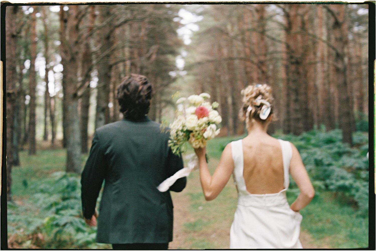 whimsical 35mm film photo of a newly married couple running in the forest with their backs to the camera, bride is holding her bouquet