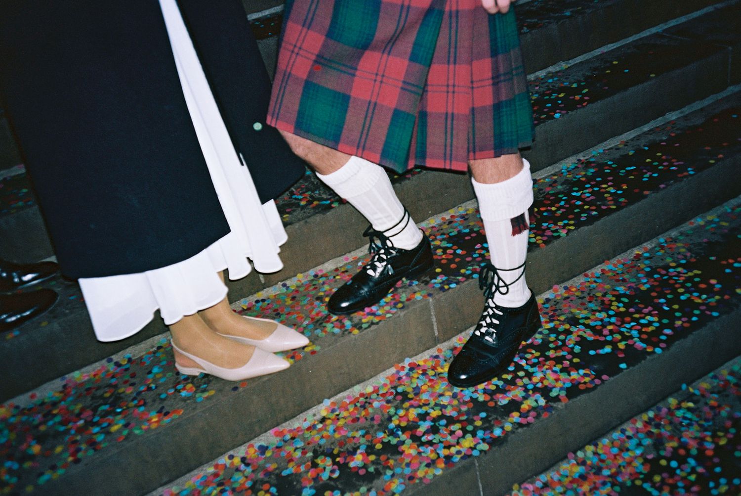 Close-up of bride and groom's feet on confetti-covered steps, the groom is in kilt and black shoes, the bride wears white flats and dress.