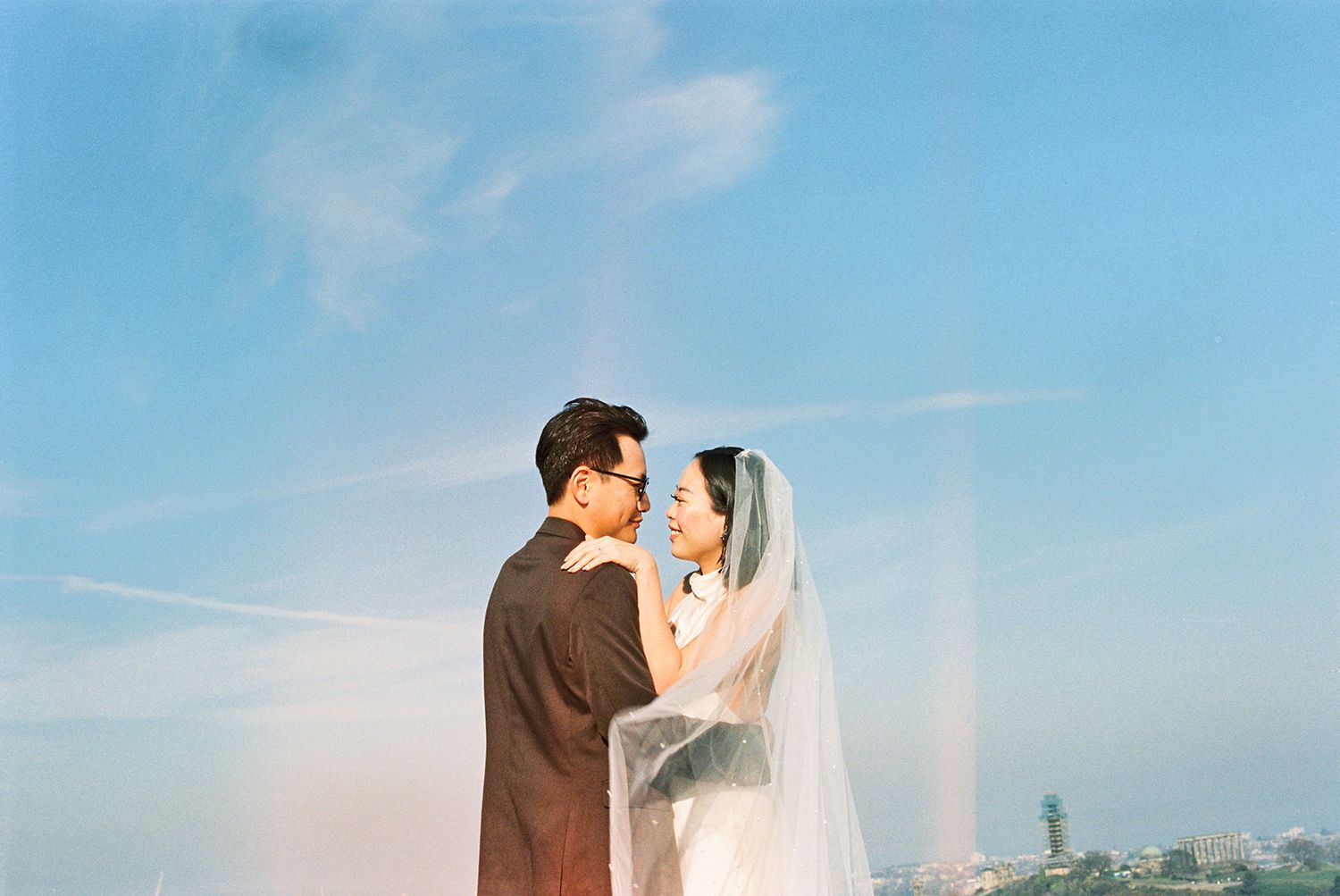 A couple in wedding attire share a romantic moment under a blue sky in Edinburgh, Scotland. There are some light leaks in the image.