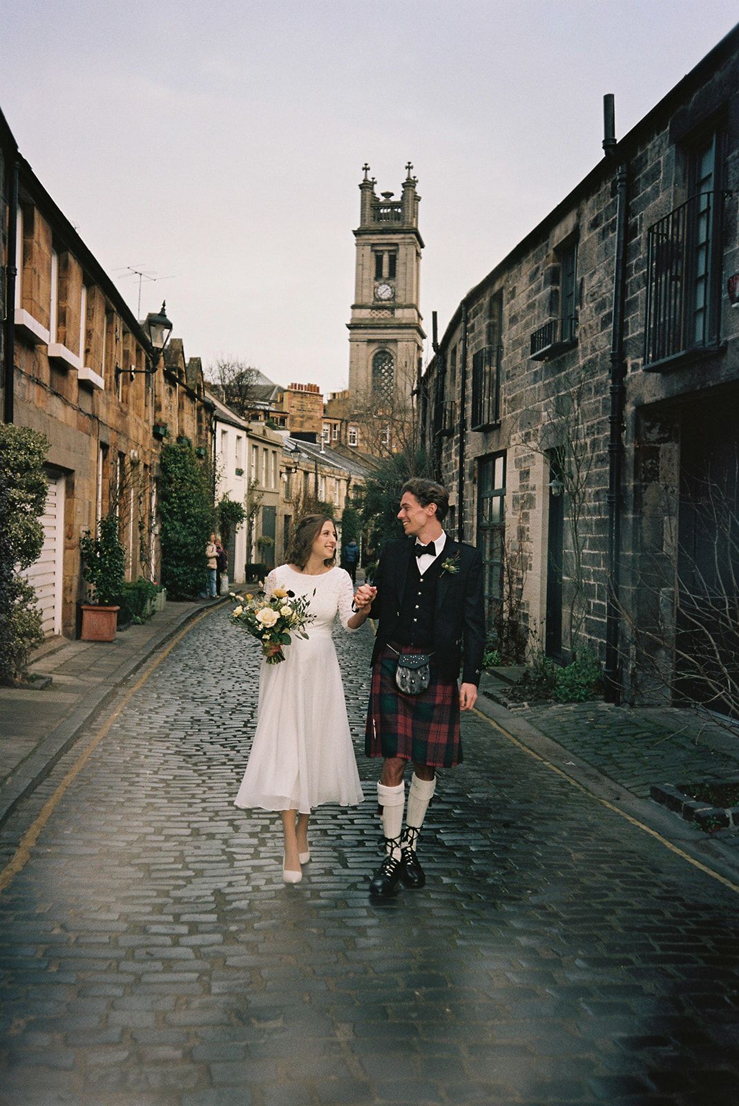 A bride and groom walk hand-in-hand on  cobblestone Circus Lane with a church tower behind them.