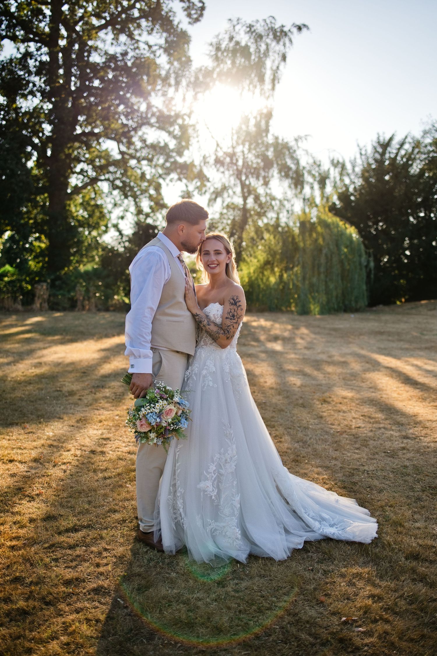 Bride and groom embrace in golden sunset light in a lush garden, bride wearing lace gown holding floral bouquet.