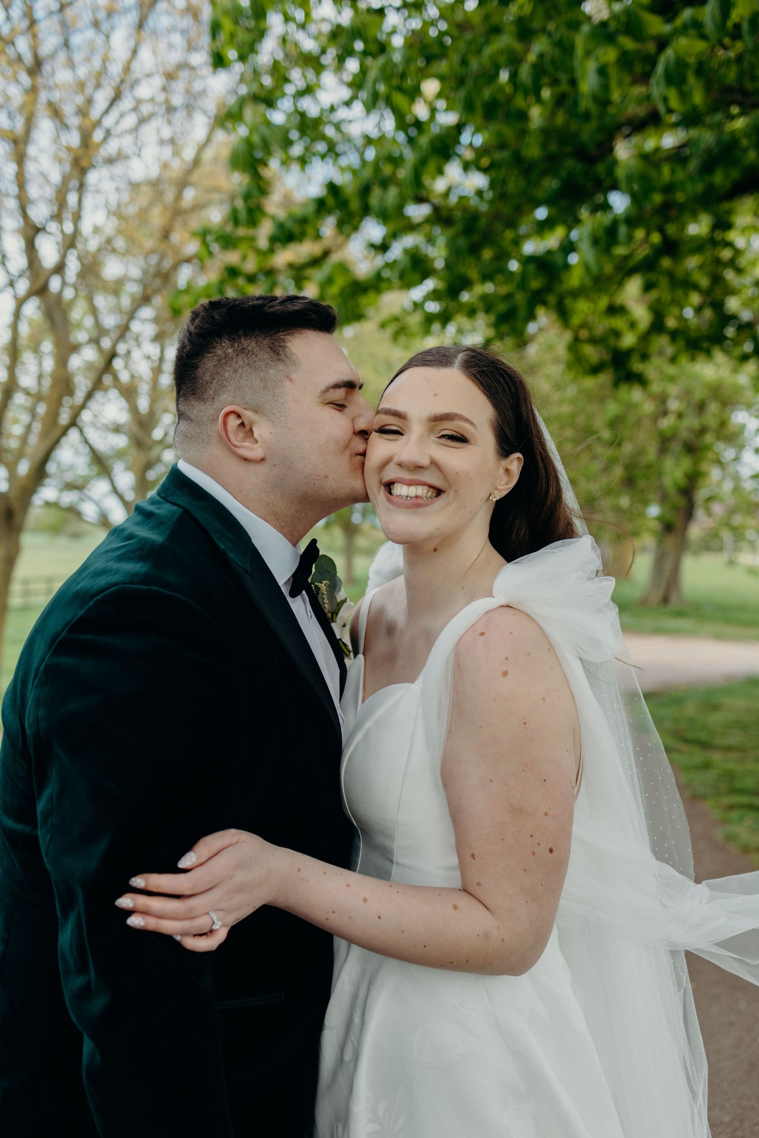 Groom in dark suit kisses bride in white gown outdoors, she smiles radiantly, surrounded by lush green trees.
