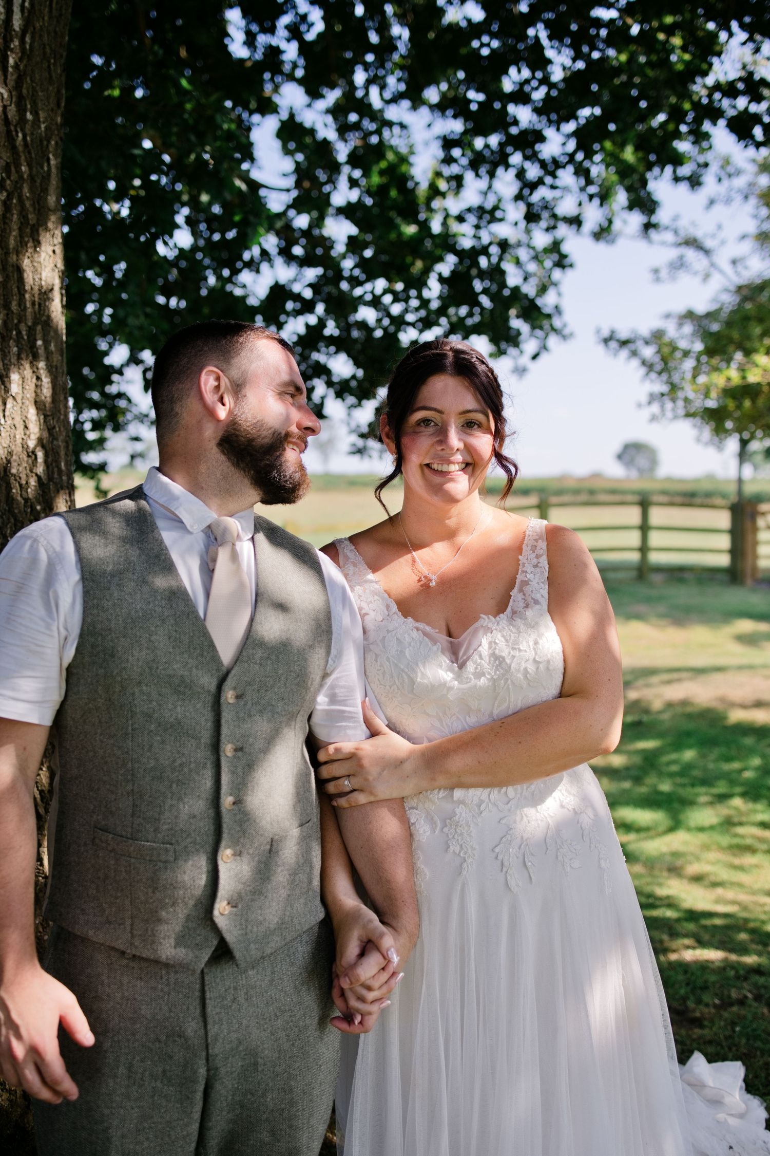 A smiling bride in lace gown holds hands with bearded groom in grey vest suit beside a tree outdoors.