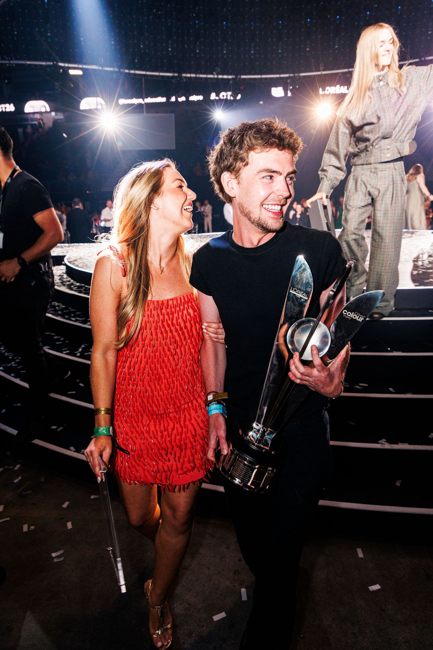 Man holding trophy poses with woman in red dress at an awards ceremony with stage lights in background.