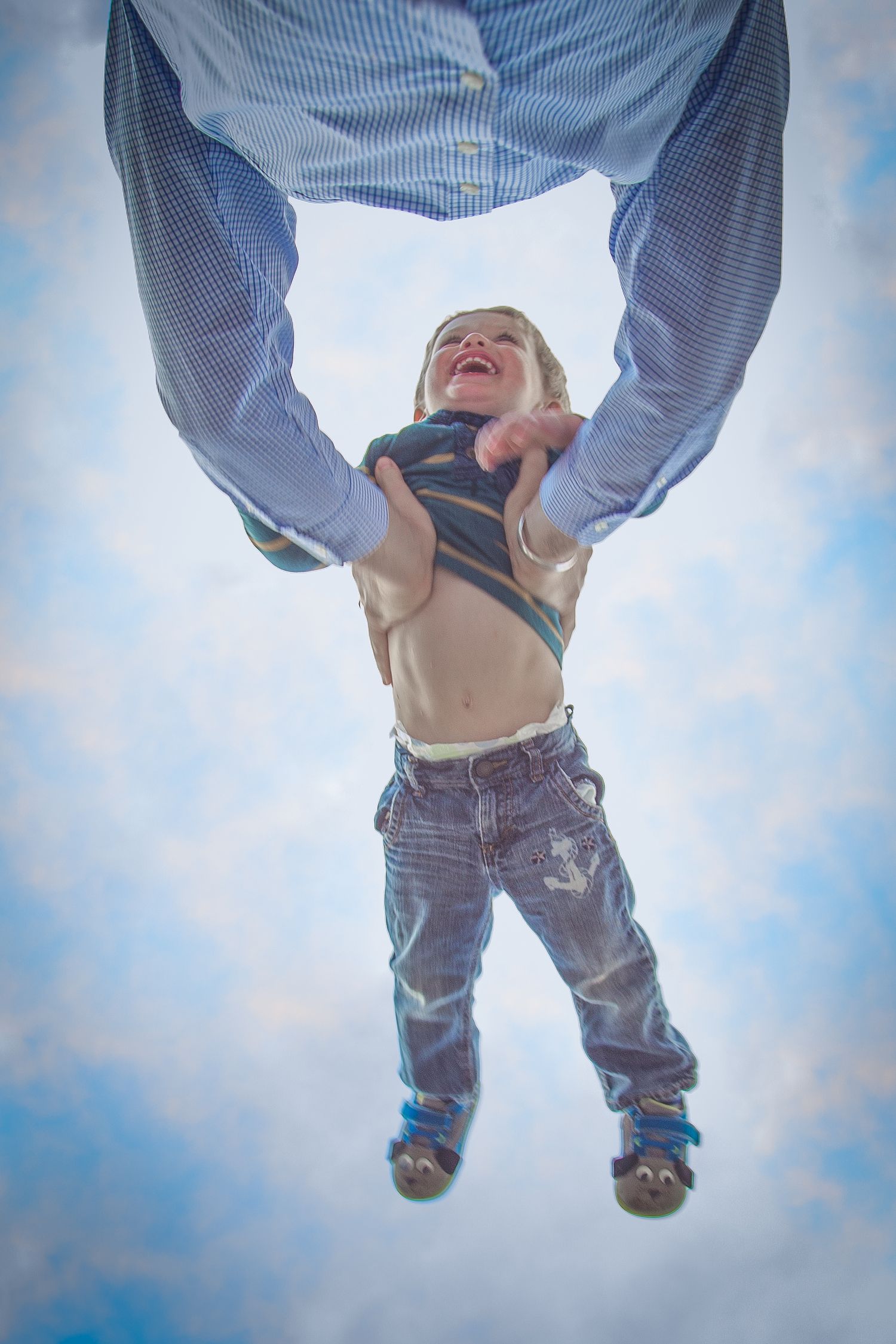Playful low-angle photo of someone lifting a child against blue sky background.