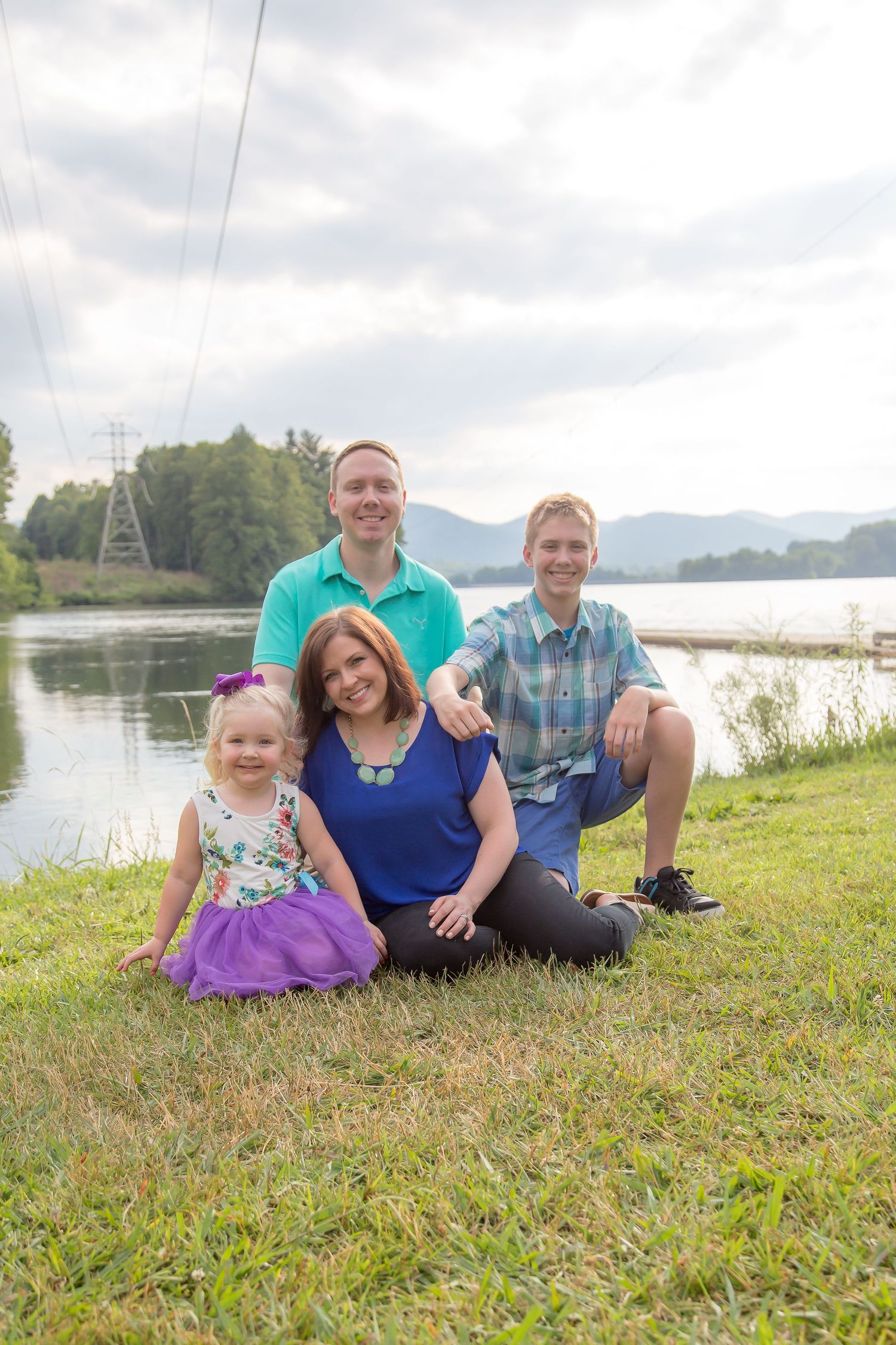 Family sits together on grass with scenic lake and mountain landscape behind them.