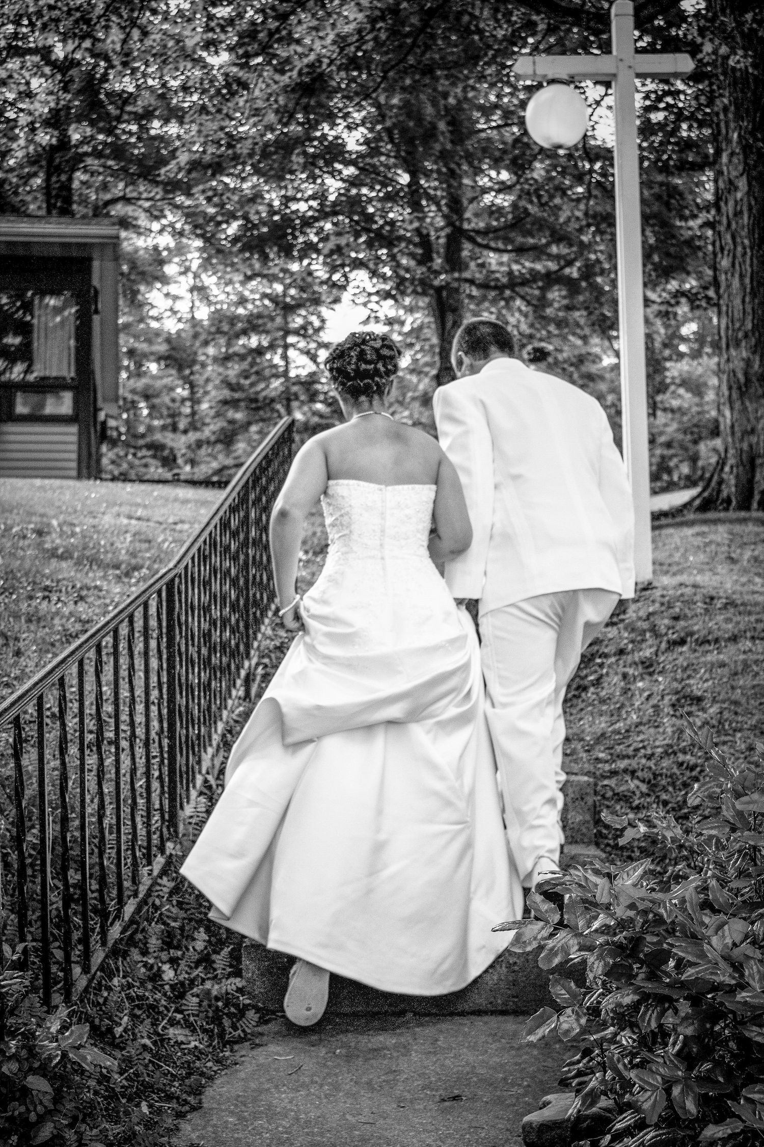 A couple in wedding attire walks down garden steps with iron railings in a black and white photograph.