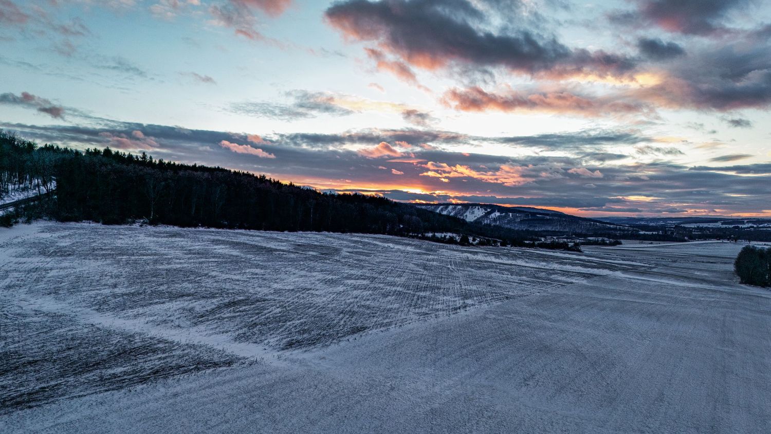 A snow-covered winter road winds through mountains under dramatic pink and purple sunset clouds at dusk.