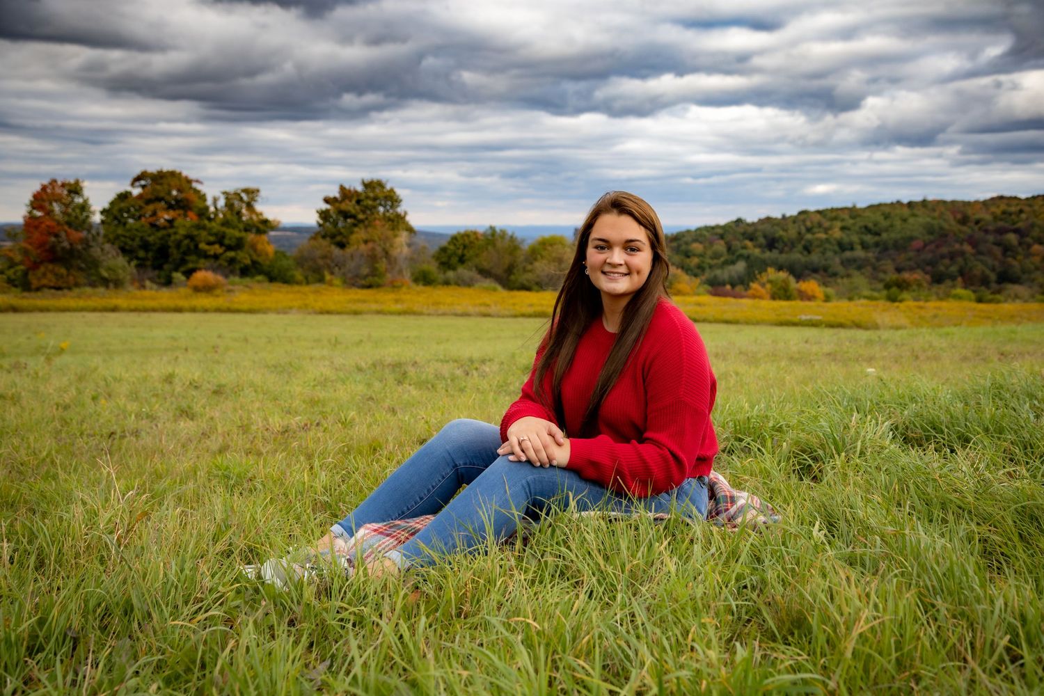 Woman in red sweater sitting in autumn field with cloudy sky and fall foliage in background.