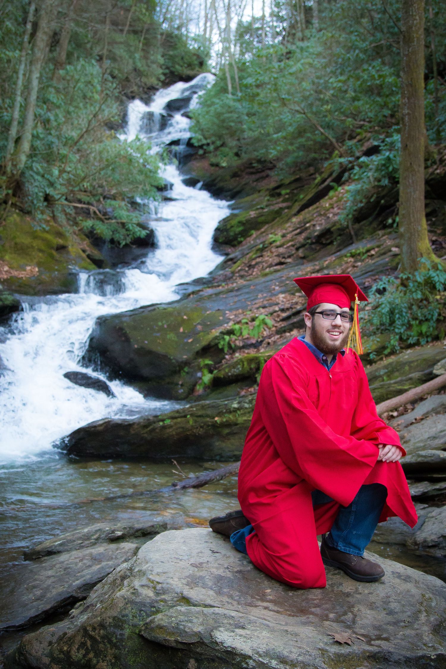 A cascading waterfall flows through a lush forest setting while a graduate in red cap and gown poses on rocks.