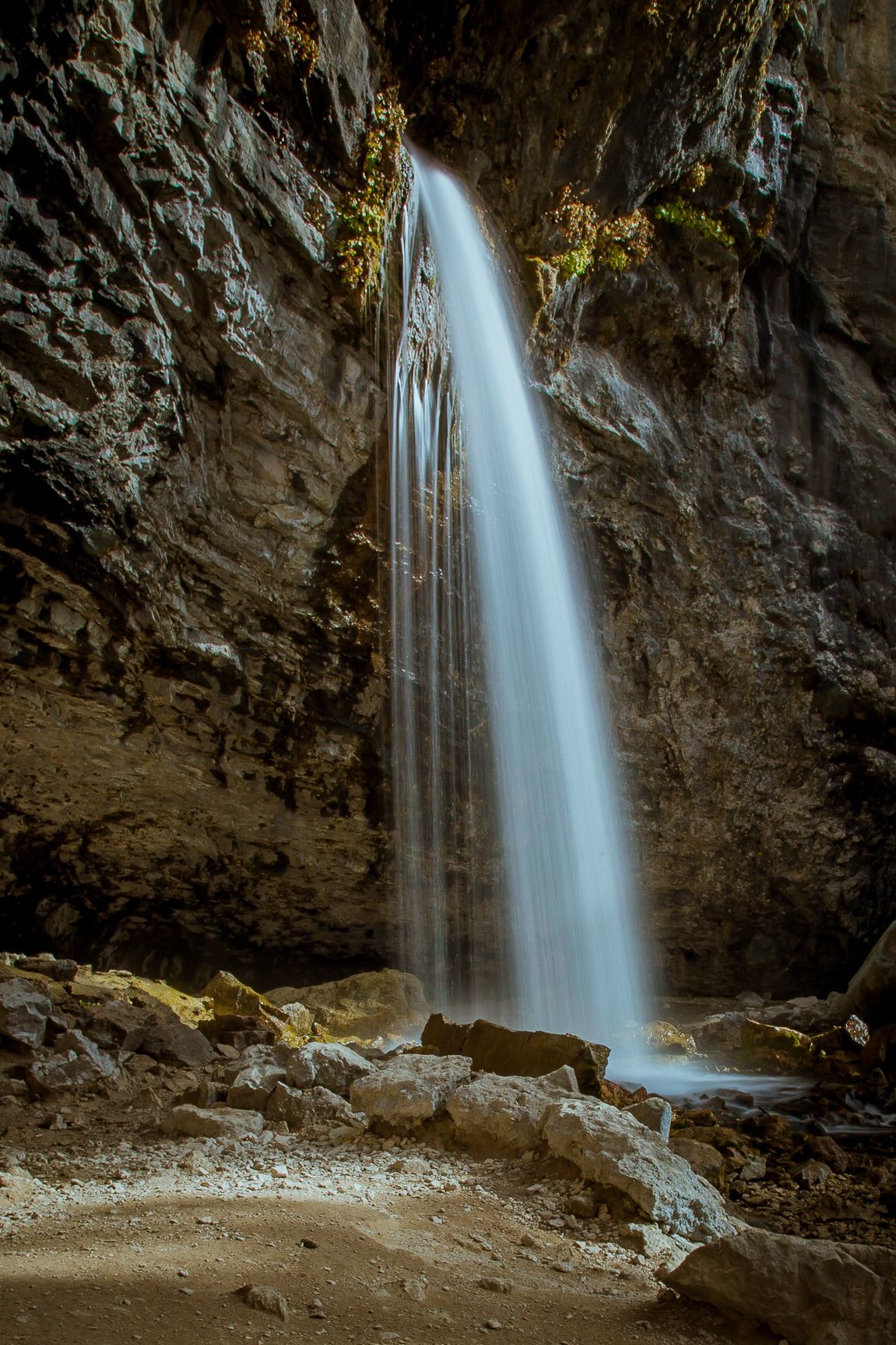A majestic waterfall cascades down rocky cliffs inside a dark cave with moss-covered walls and natural ice formations.