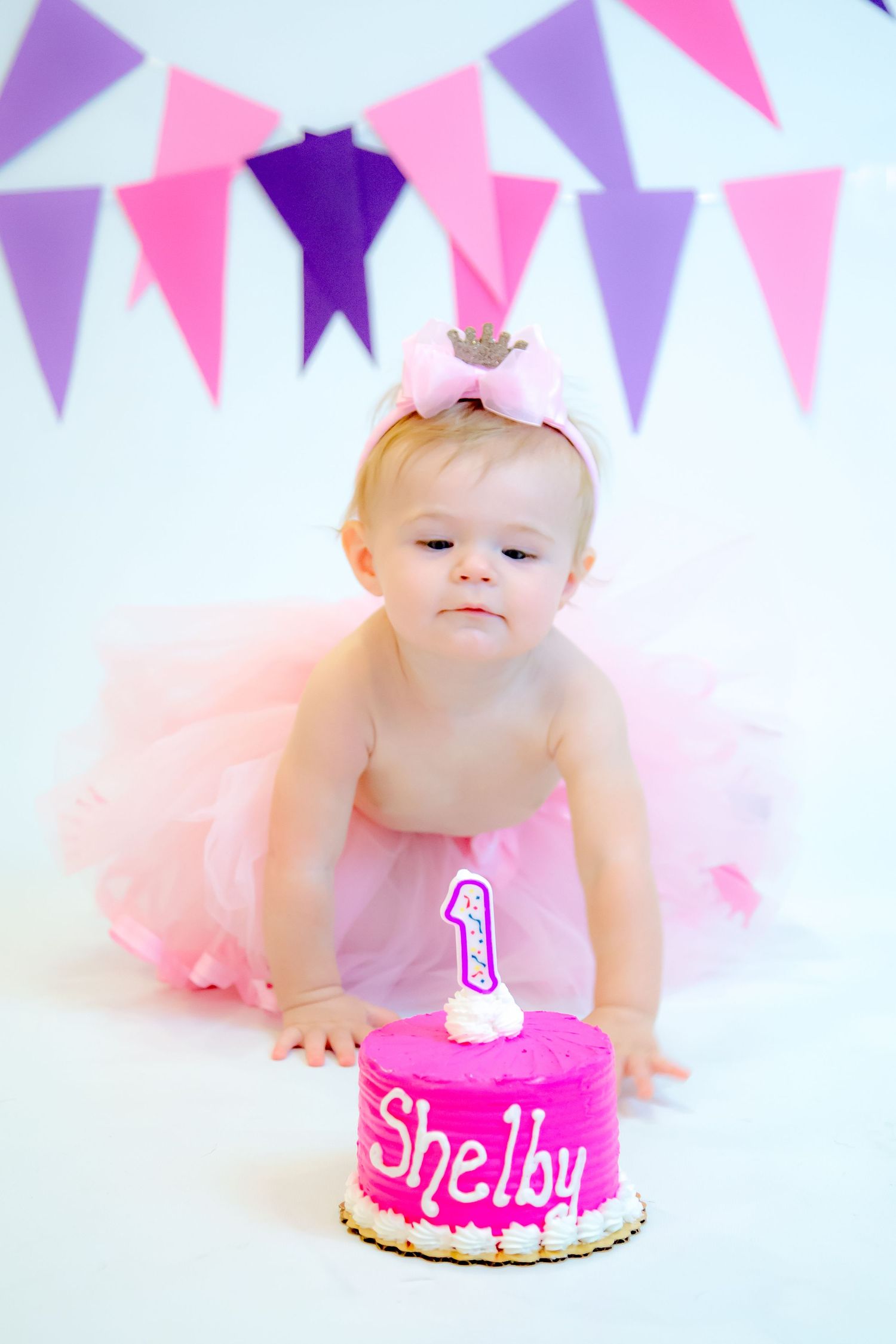 First birthday celebration with pink cake and tutu against purple and pink bunting banner backdrop.
