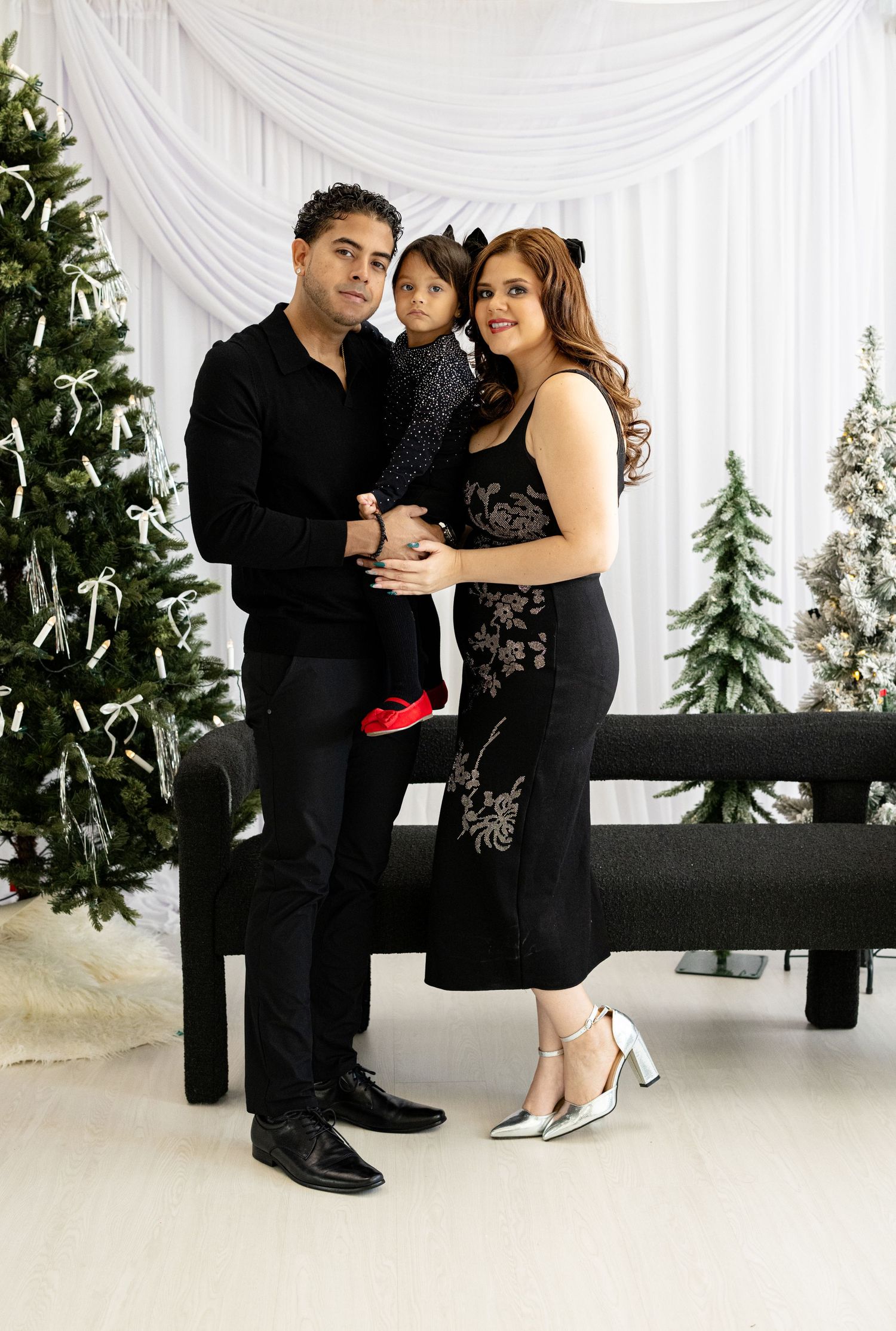 Elegant family portrait in formal attire against white draped backdrop with Christmas trees.