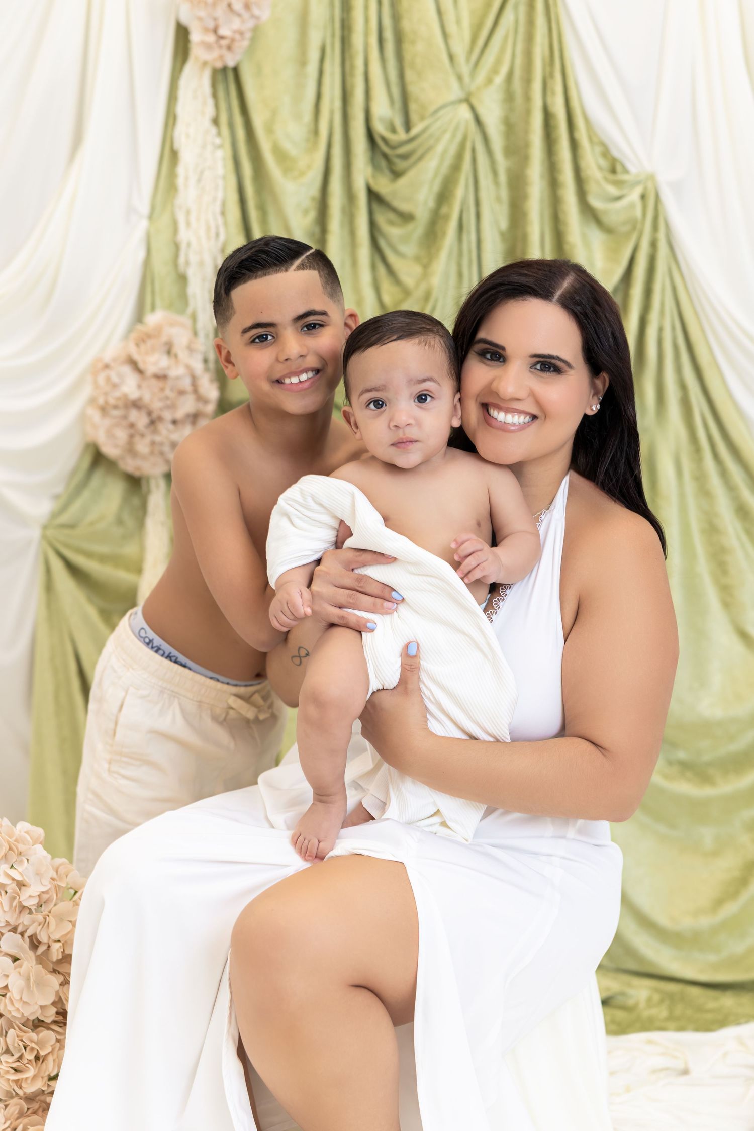 A smiling woman in white dress holds a baby while a shirtless boy poses beside them in a green draped studio.