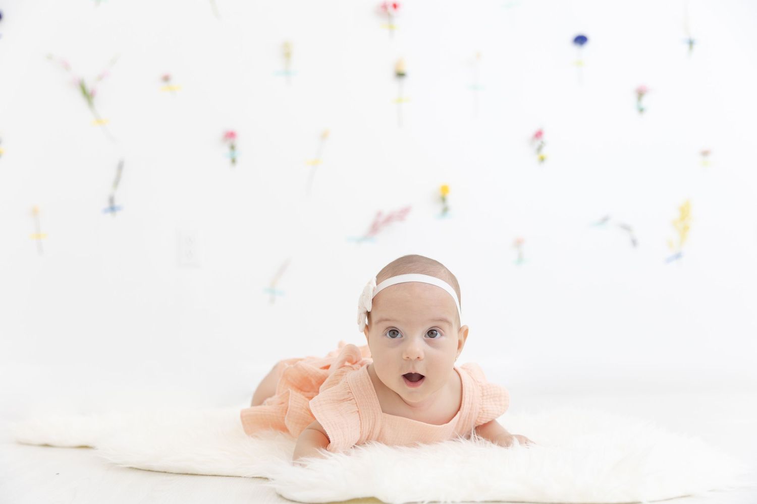 Baby in pale outfit lying on white floor with colorful confetti scattered around.