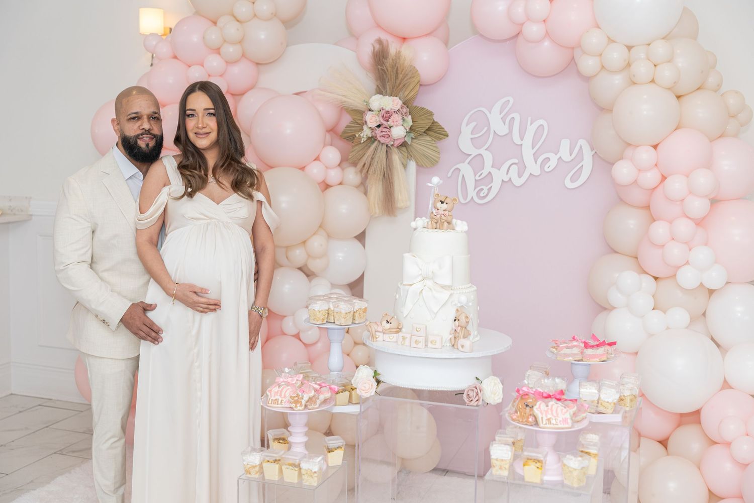 Baby shower celebration with pink balloon arch, white cake display and pastel decorations.