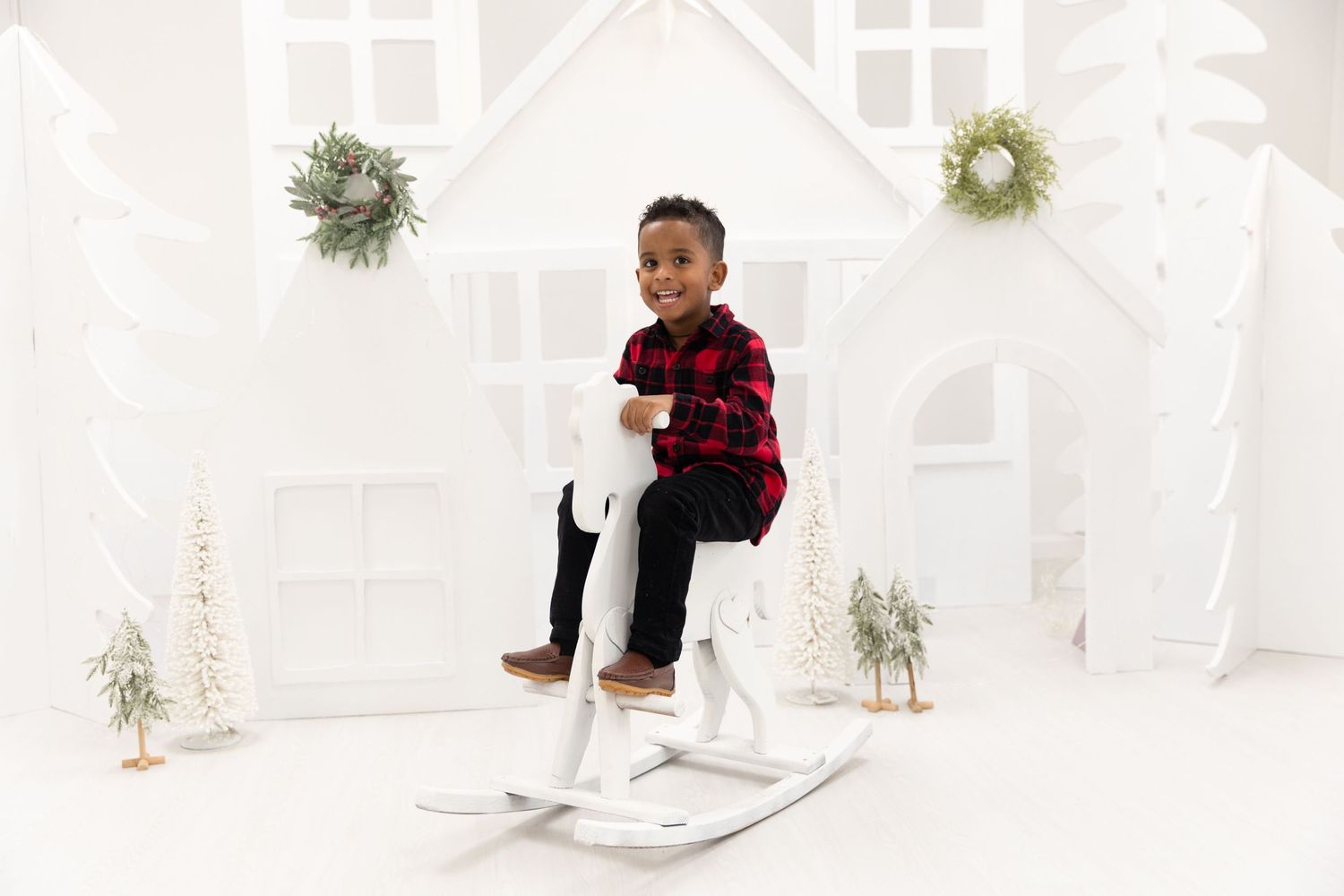 Child in buffalo plaid shirt sitting on white rocking horse with holiday wreaths in background.