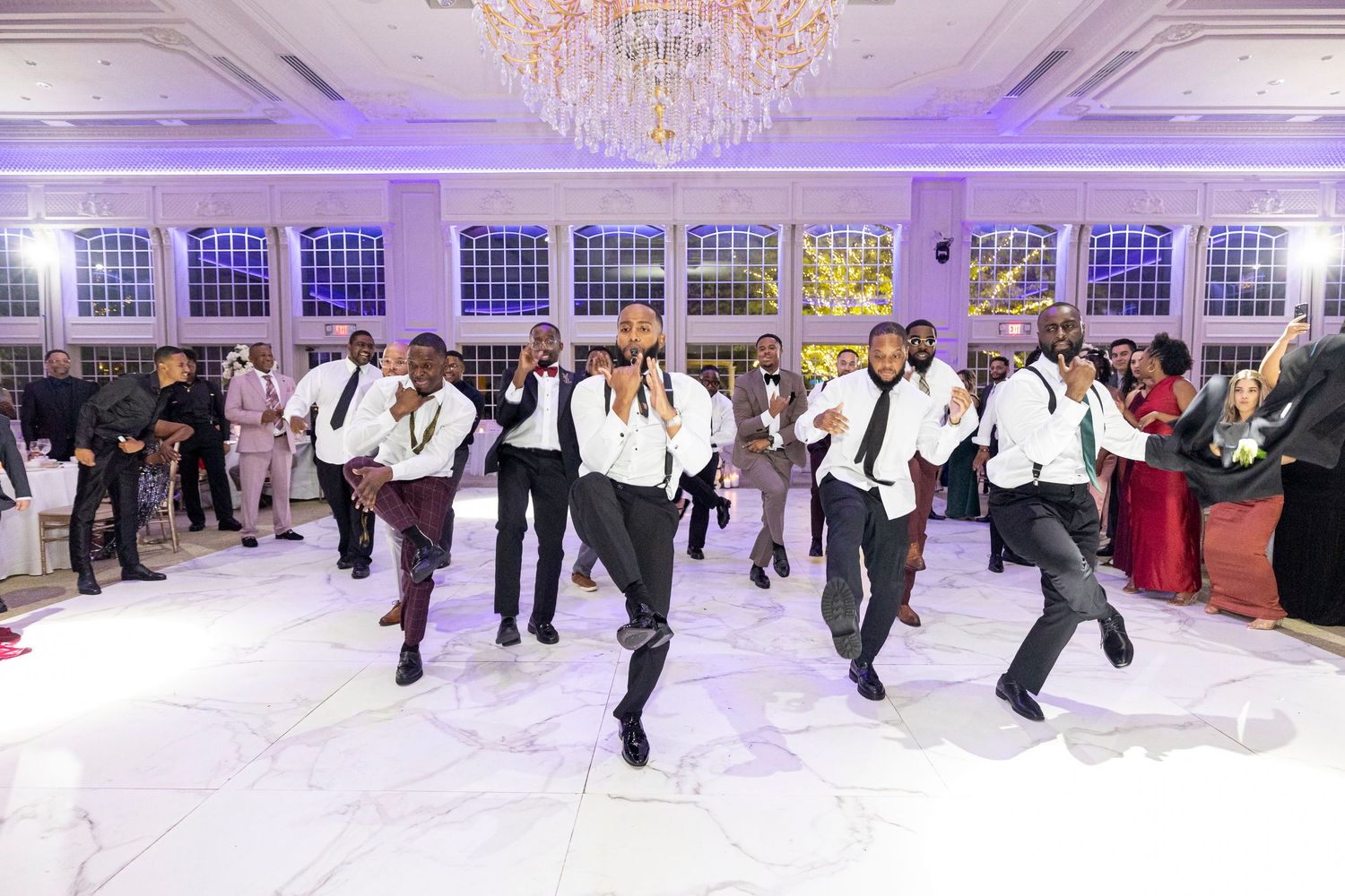 Groomsmen perform choreographed dance routine on white dance floor in elegant ballroom with crystal chandeliers.