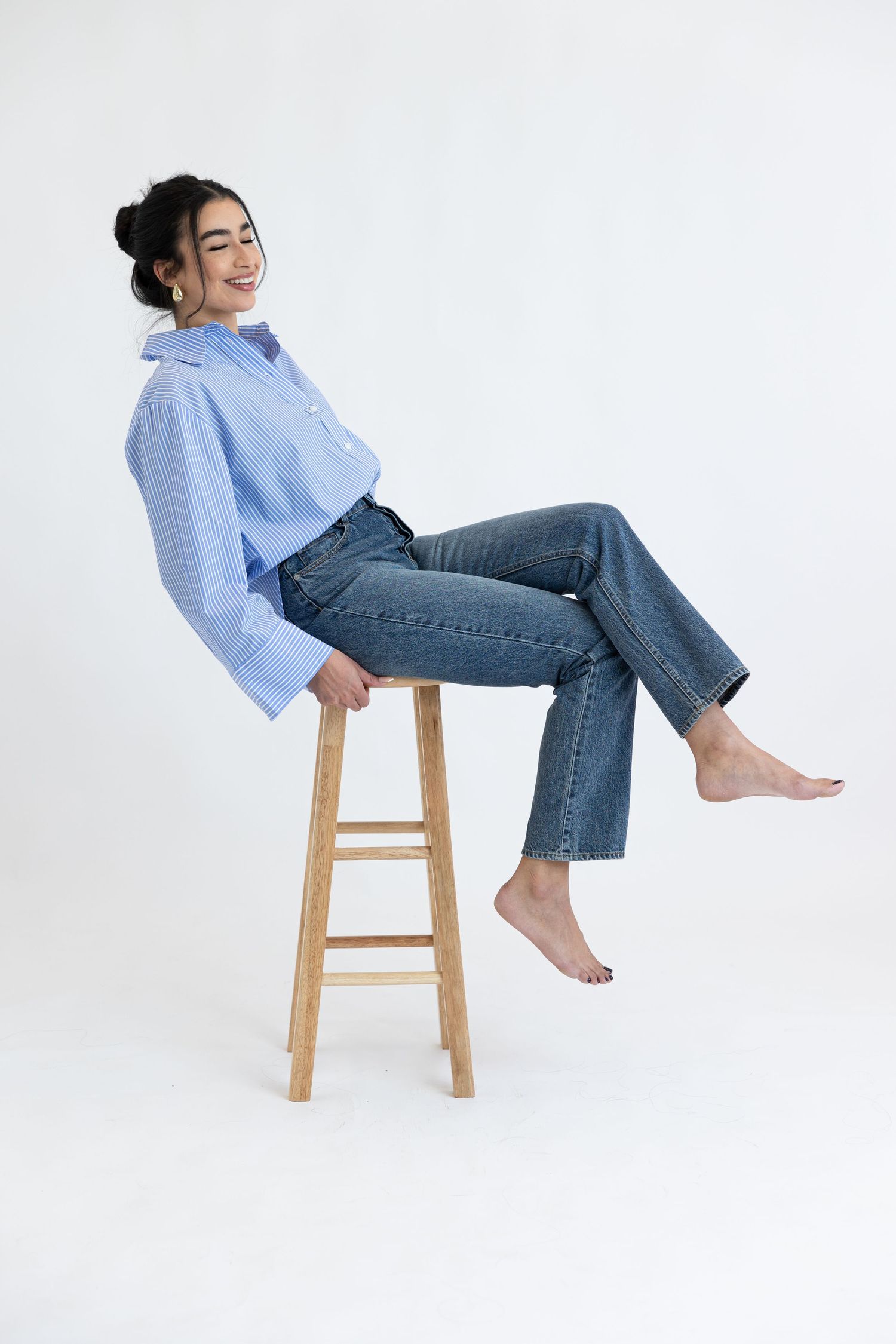 Casual portrait of woman in blue shirt and jeans sitting barefoot on wooden stool.