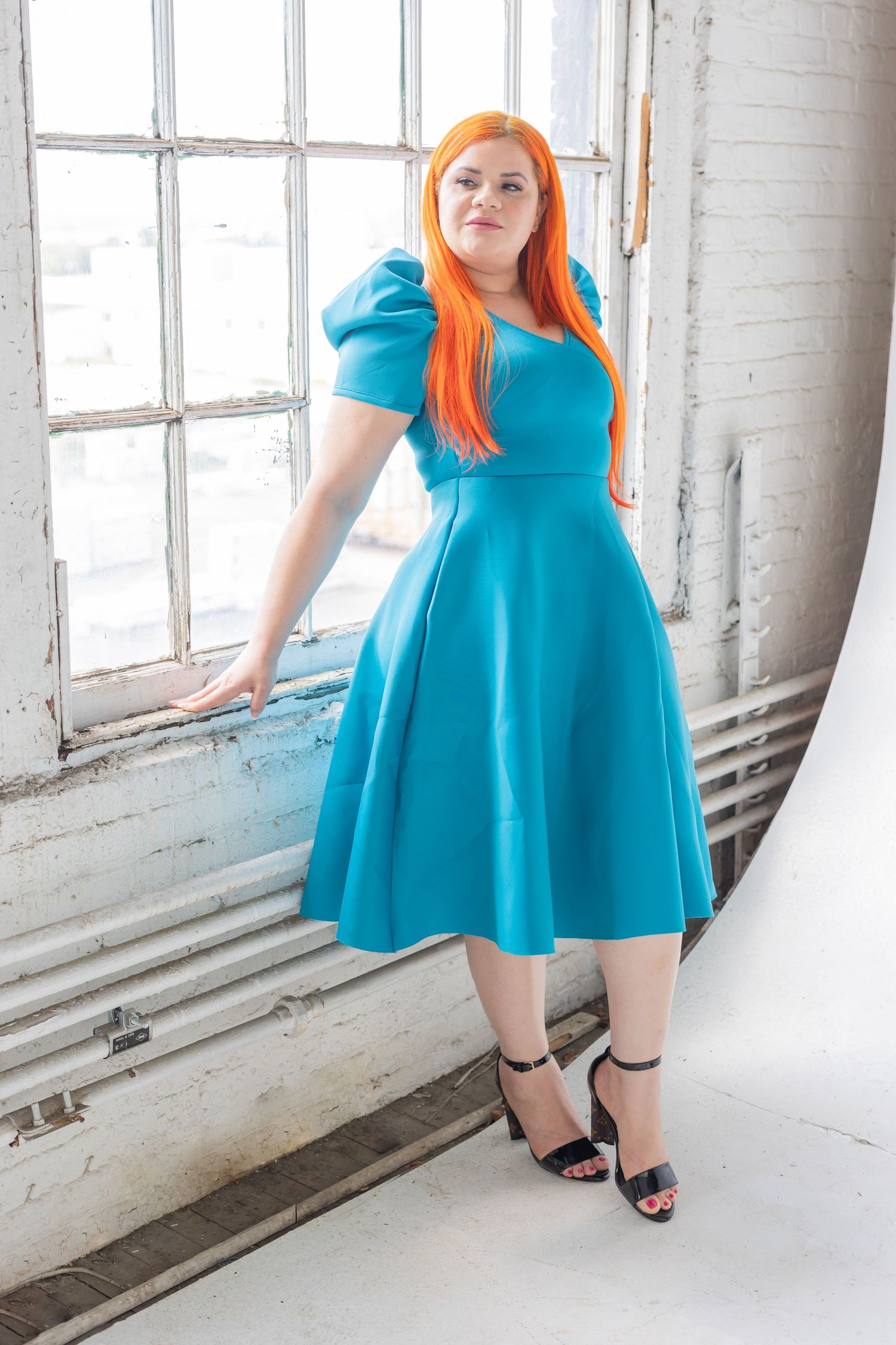 Woman in vibrant turquoise midi dress with bright orange hair poses against white industrial window backdrop.