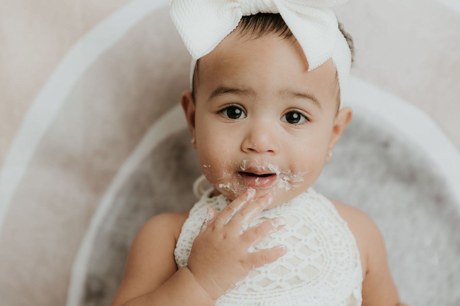 Baby in white bow headband sitting in tub with frosting on face.