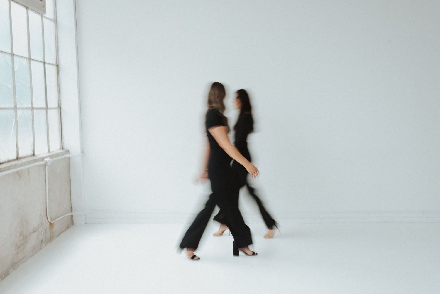 Two people in black outfits dance together against a white wall with large windows creating a minimalist aesthetic.