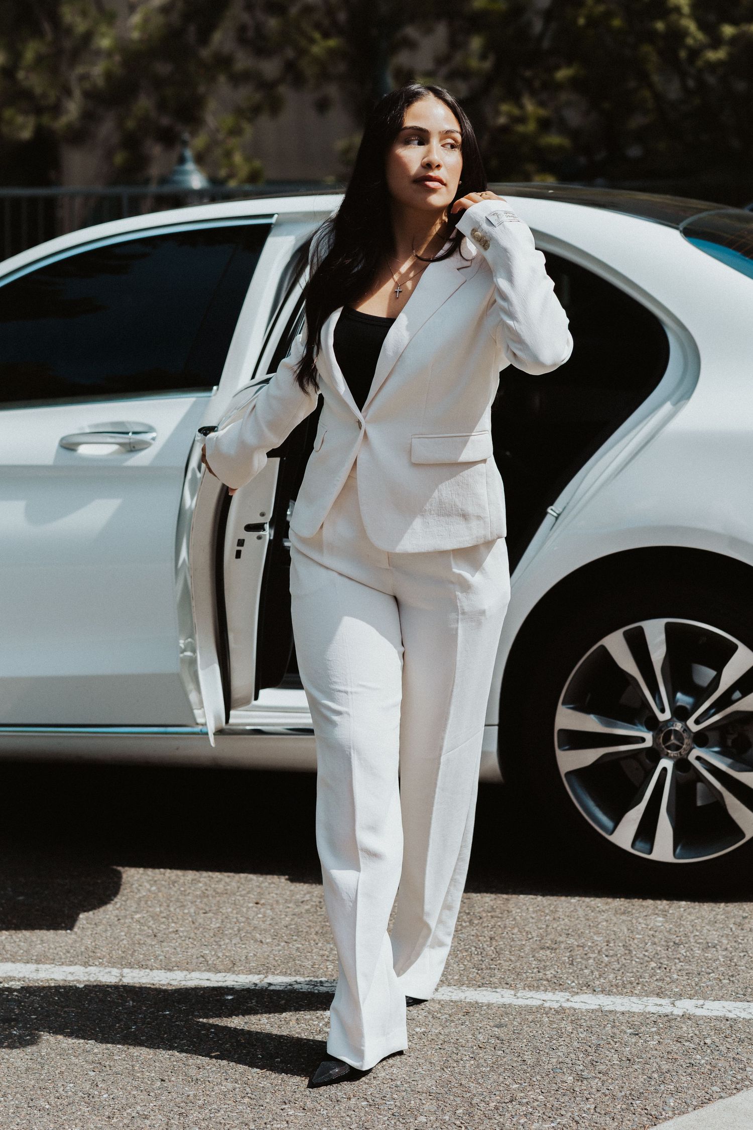 Woman in crisp white pantsuit walking confidently next to a luxury car.