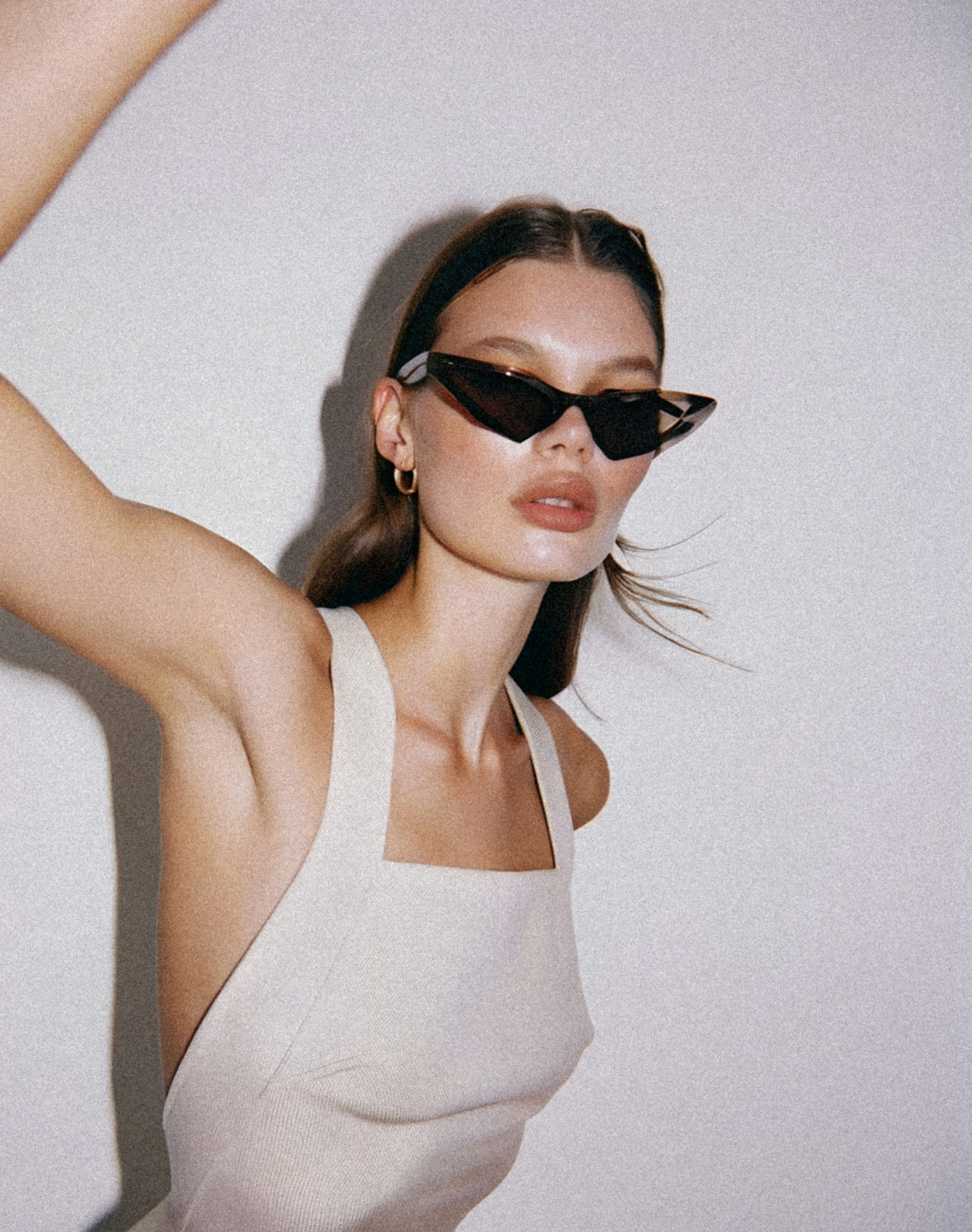 Woman in white tank top wearing trendy cat-eye sunglasses, posing with arm raised against a neutral background.