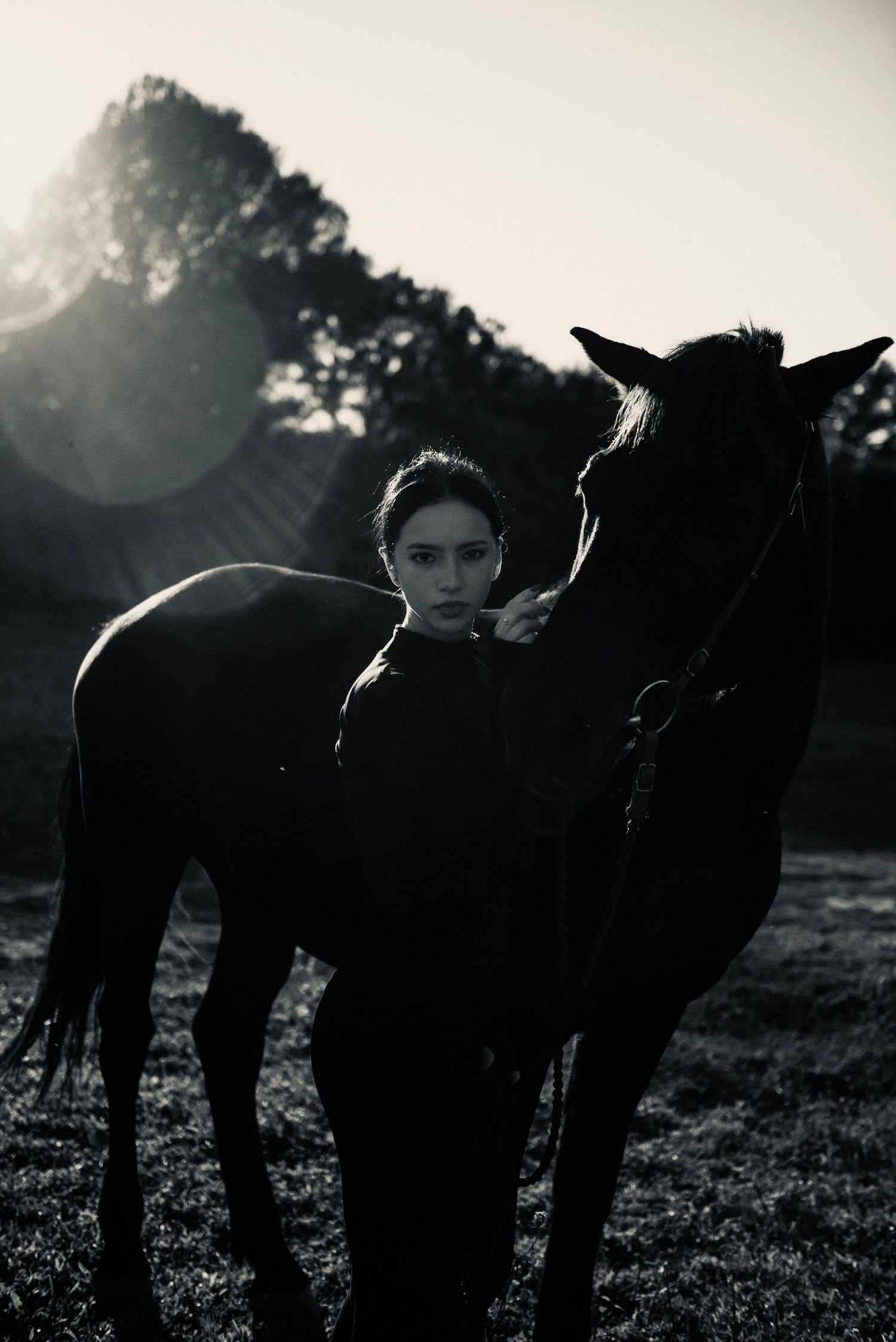 Black and white photo of a young woman standing beside a dark horse in a sunlit field with trees in background.