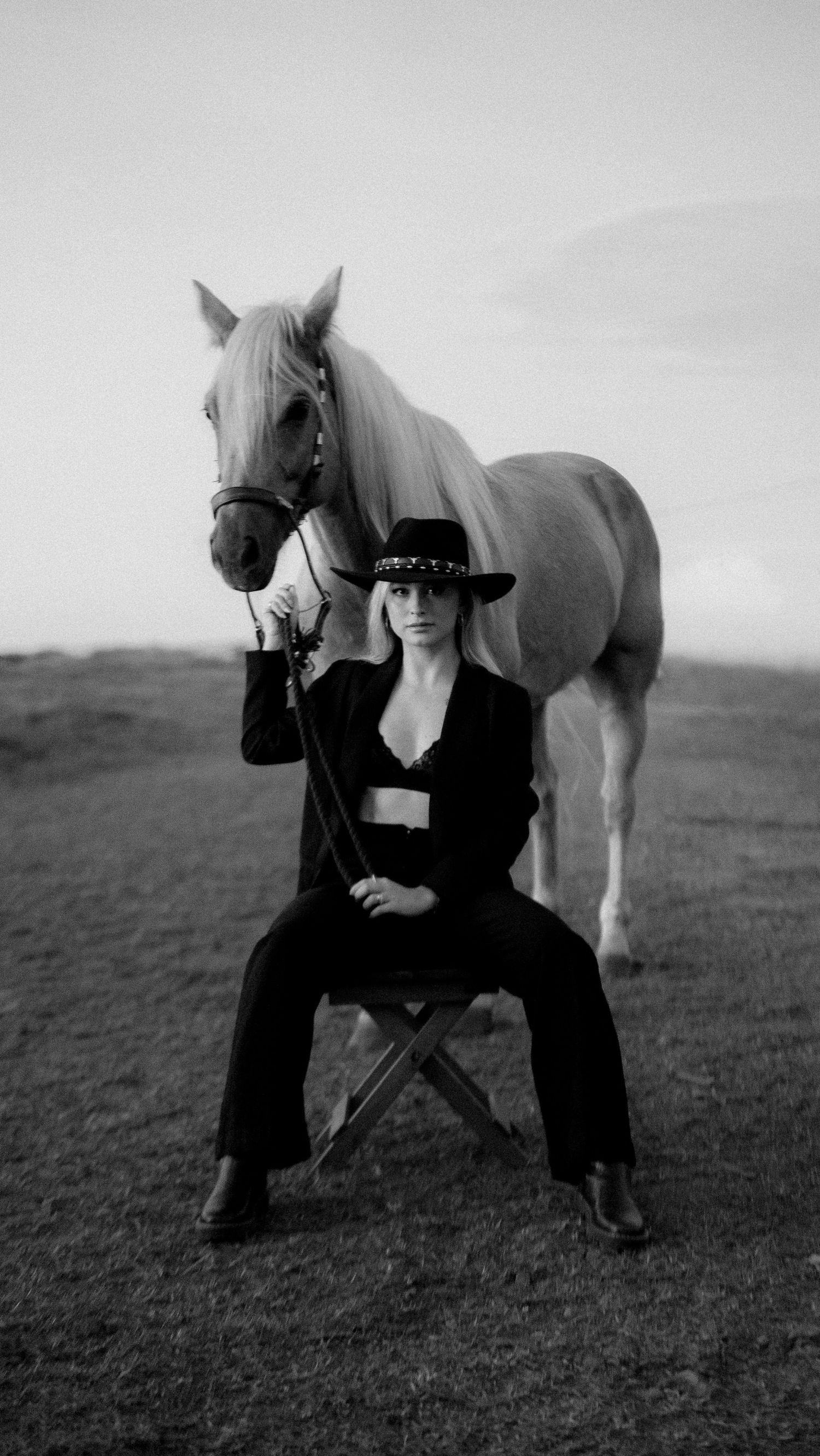 Black and white photo of a woman in cowboy hat sitting before a white horse in an open field.