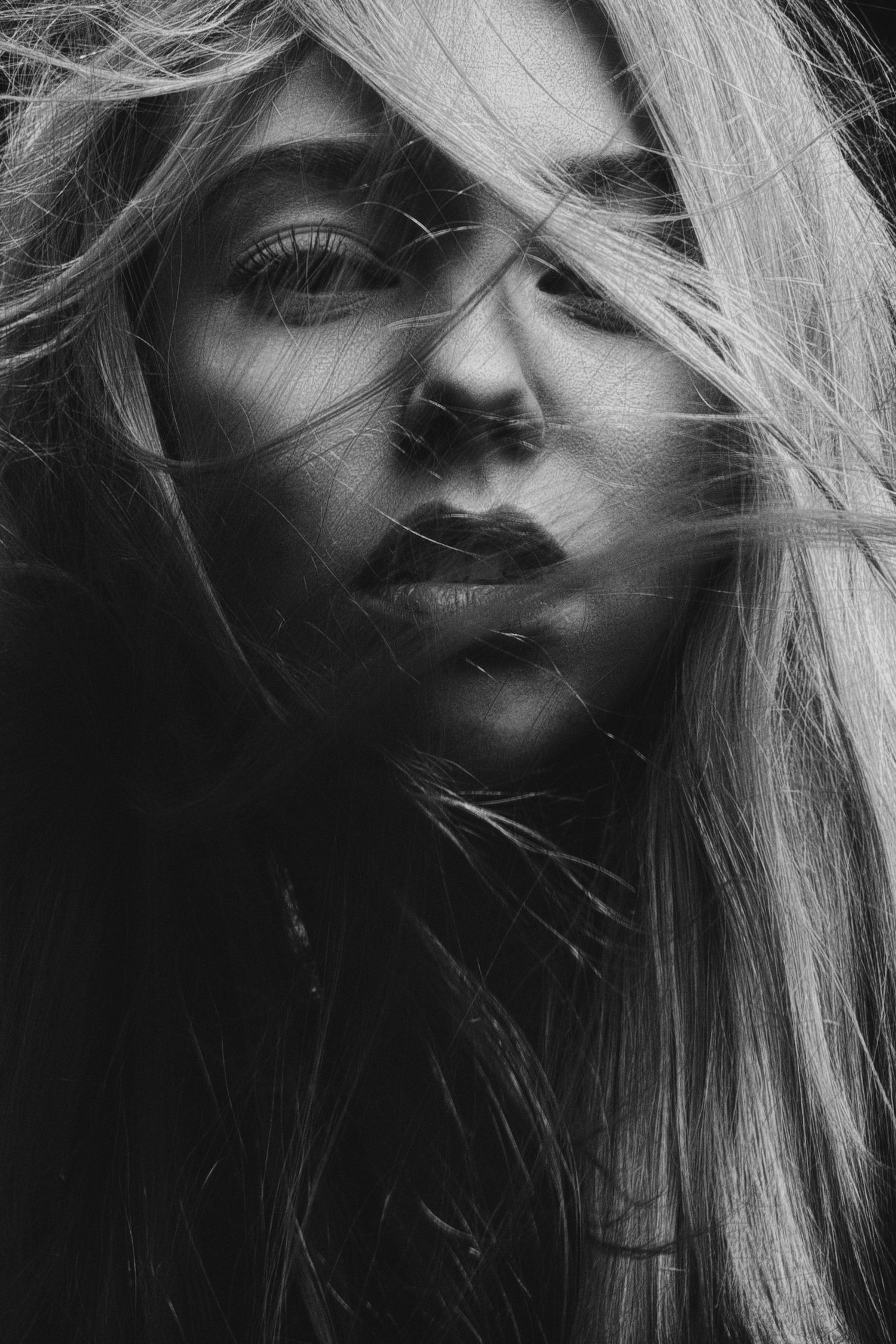 Black and white portrait of a woman with long blonde hair blowing across her face in a dramatic, moody close-up shot.