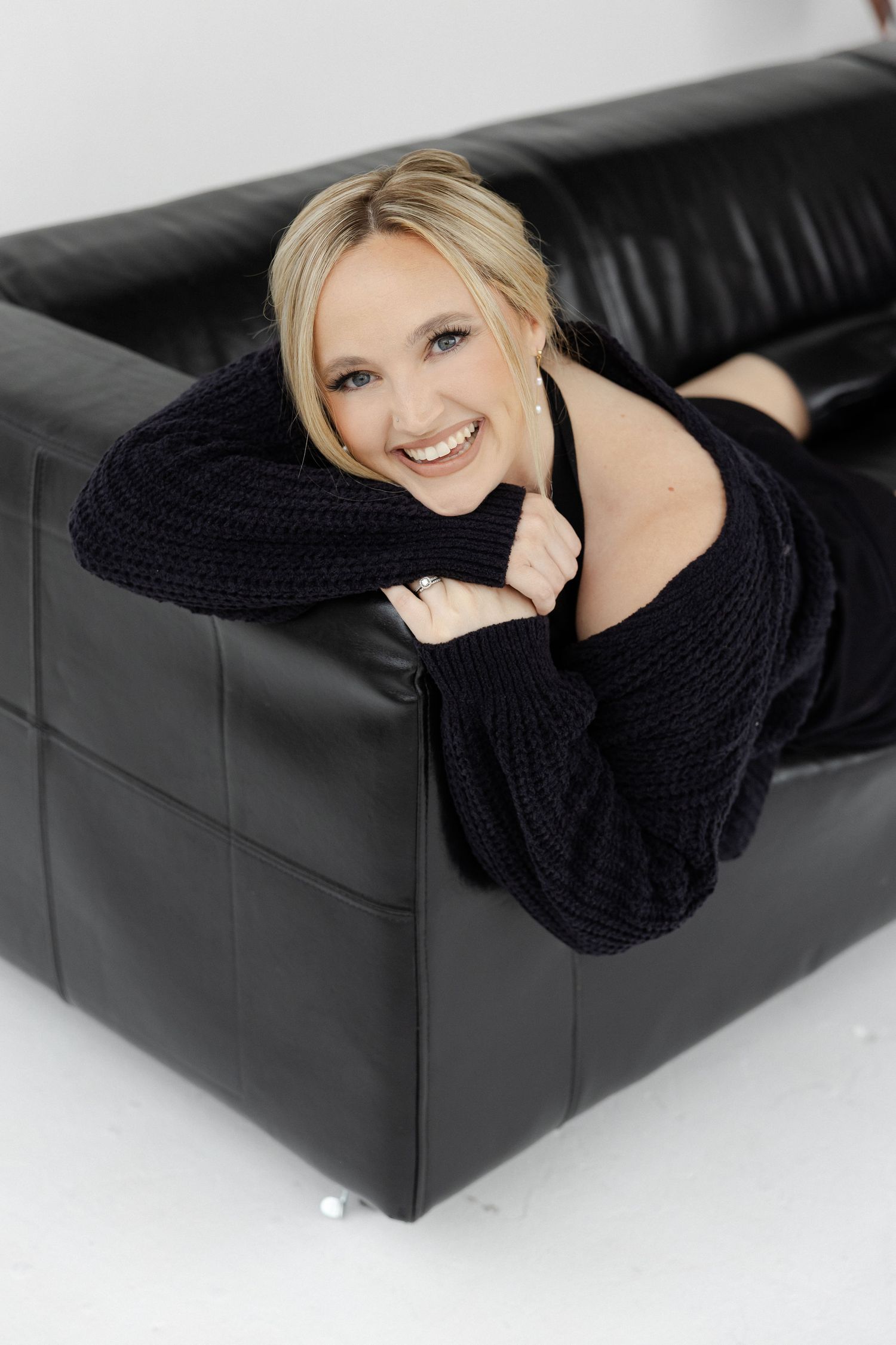 Person in black sweater lounging on dark leather couch in studio setting.