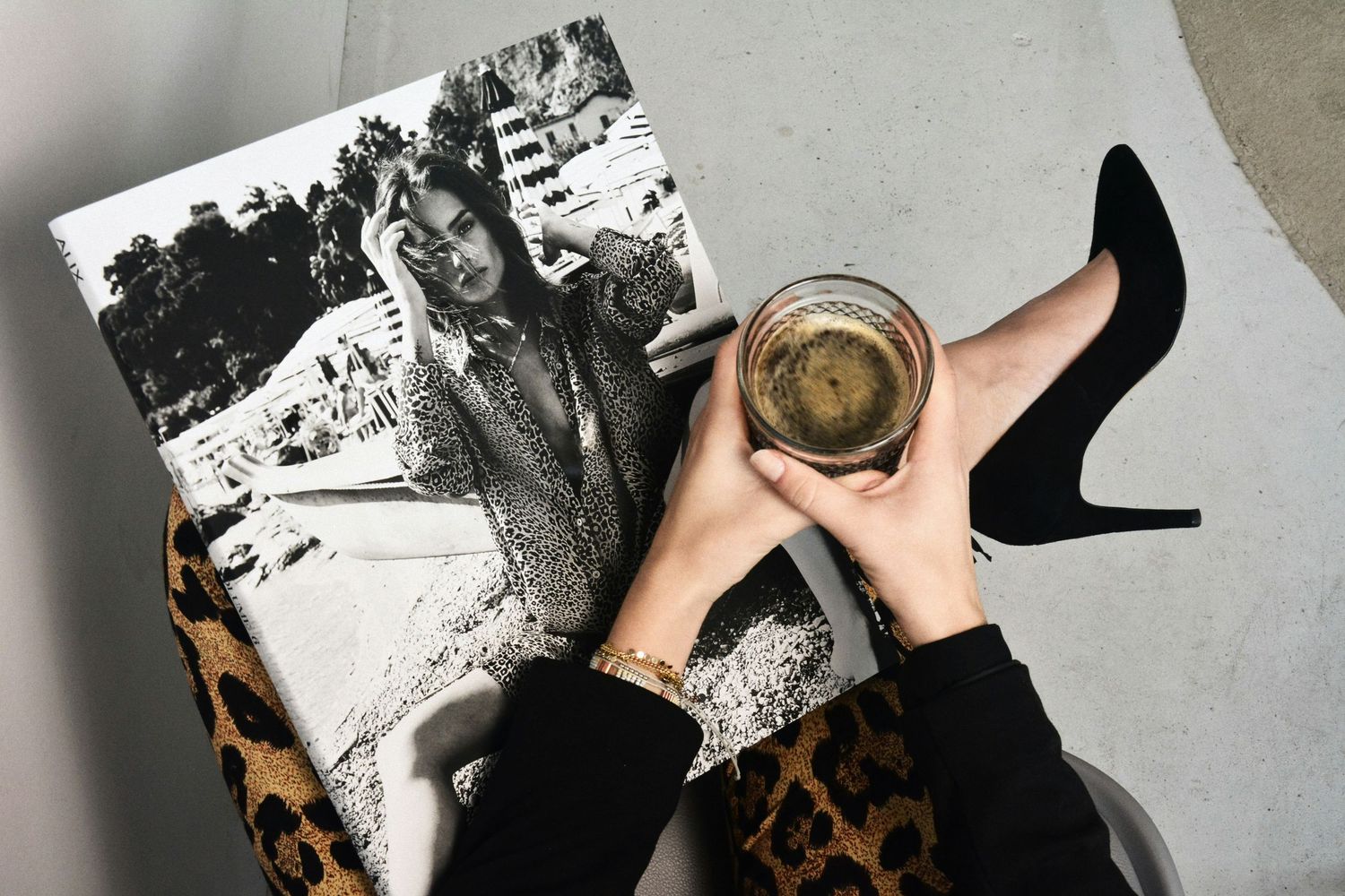 A woman holds a glass of wine beside a black and white fashion magazine, with a black stiletto heel nearby.