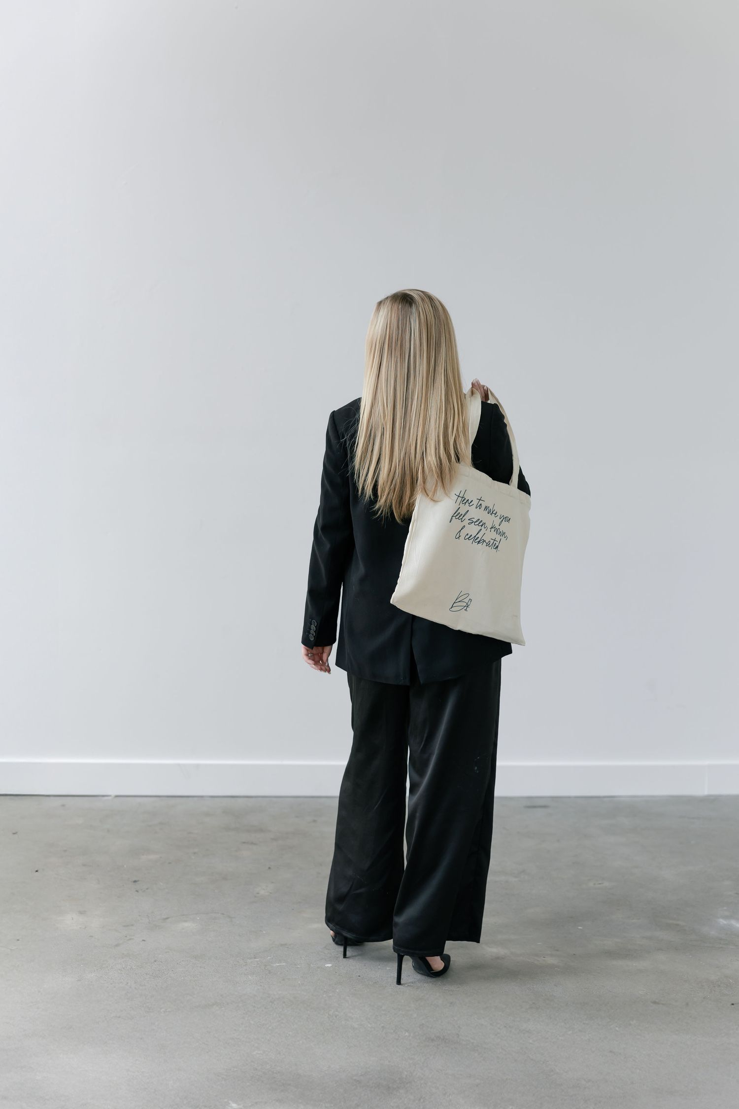 A figure in black clothing carries a canvas tote bag while walking through a minimalist white space.