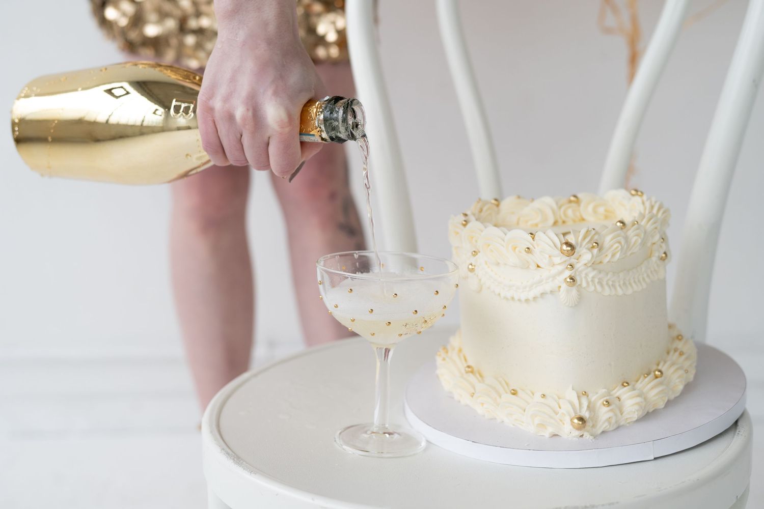 A hand pours champagne next to an elegant white cake decorated with delicate flowers on a white pedestal.