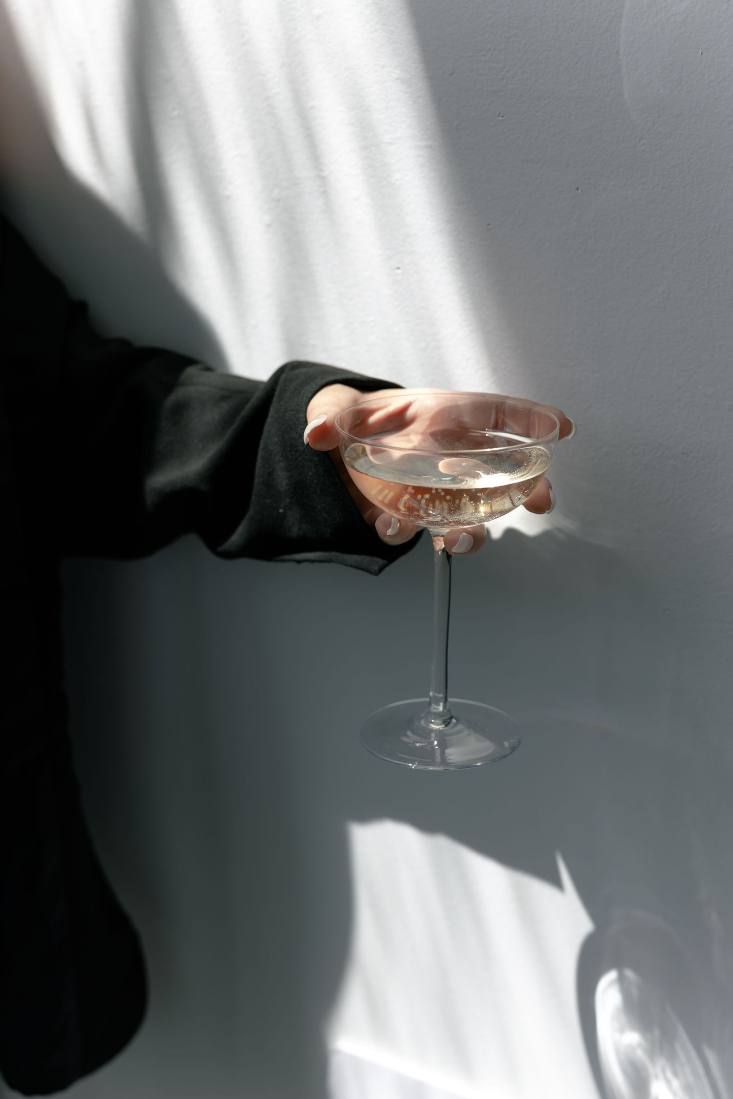 Hands holding a champagne glass against a stark white wall with dramatic shadows.