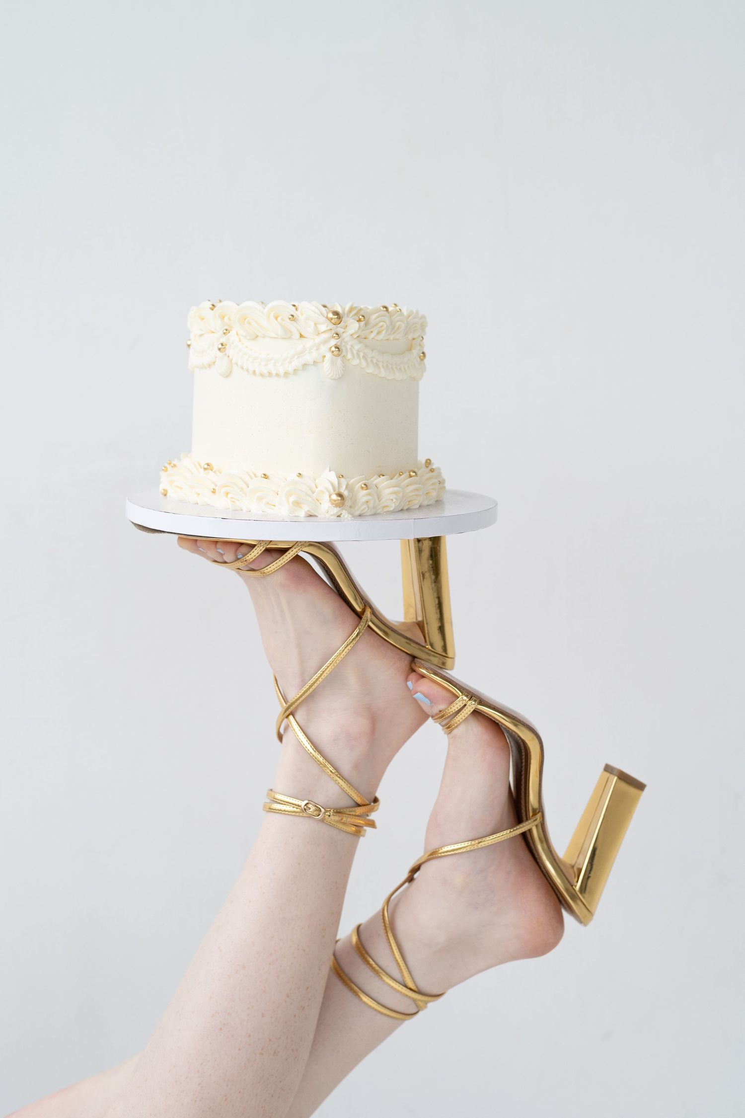 White frosted cake balanced on gold metallic high heel shoe against light background.